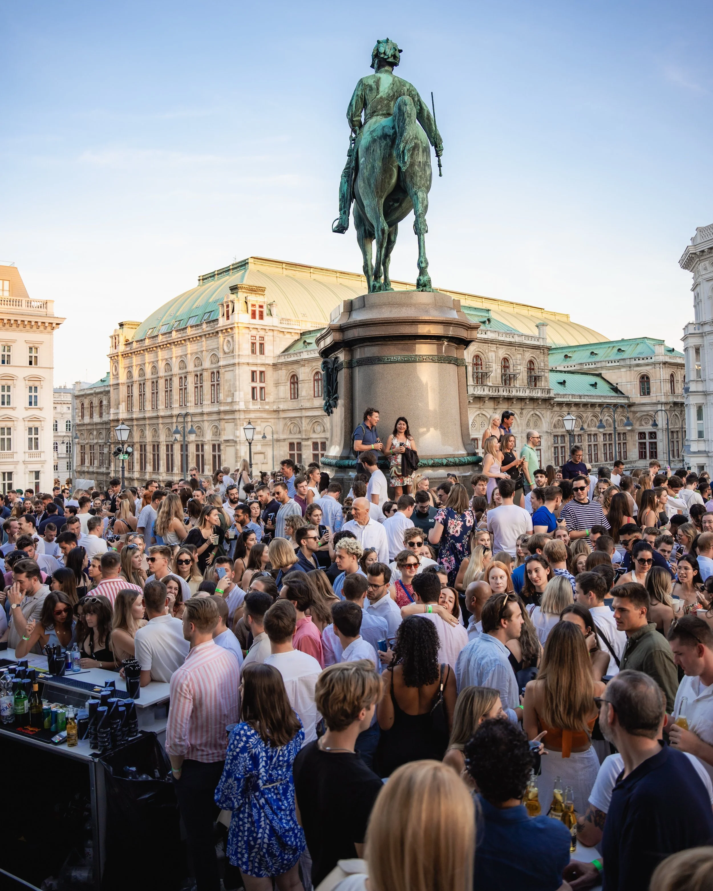 Crowd of people gathered outdoors near a bronze equestrian statue of a man. The background features historic buildings with green rooftops and a clear blue sky.