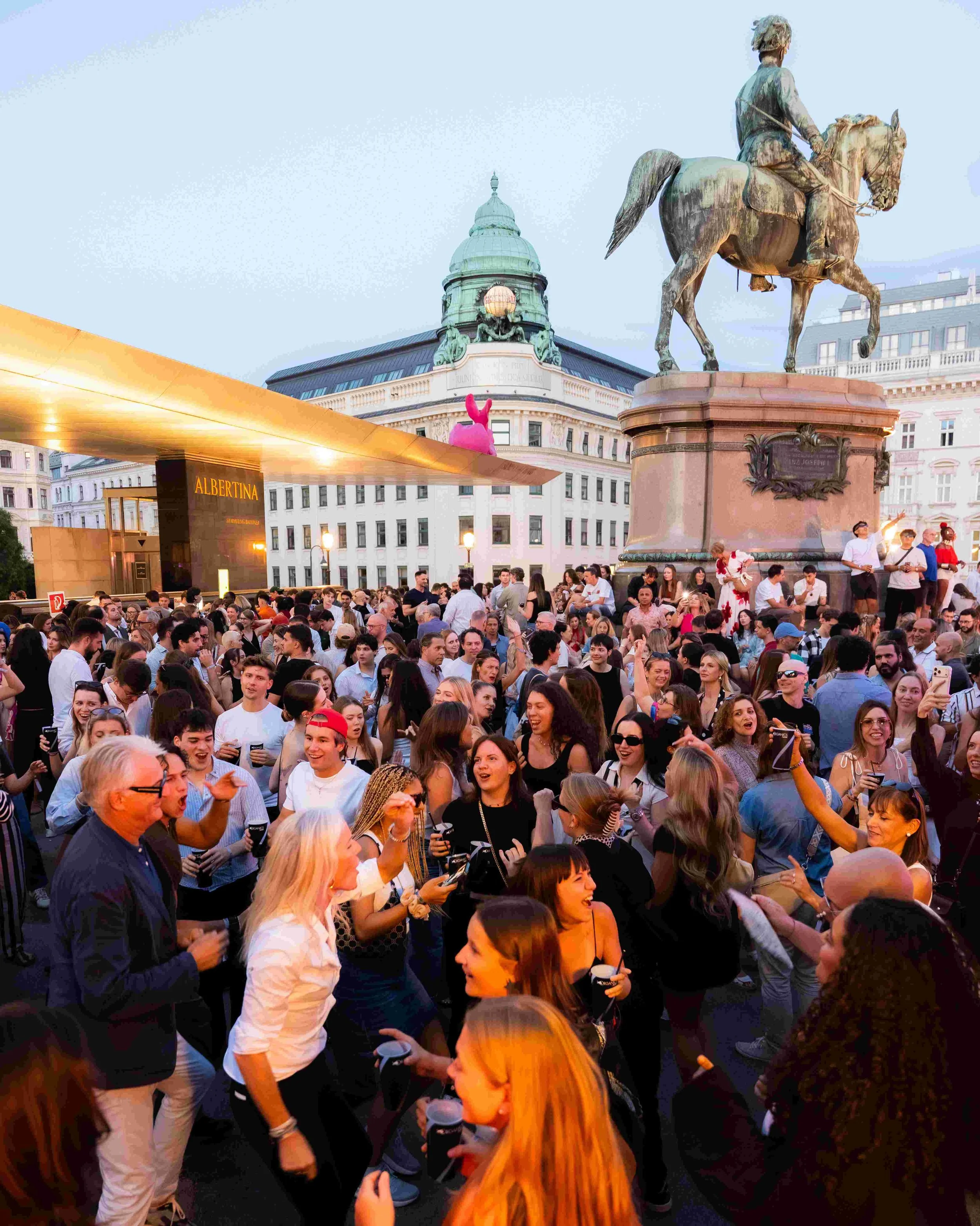 Crowd of people gathering at an outdoor event in front of a historic building with a dome and statue of a man on a horse. A pink rabbit costume is visible on a person's back near the center. The atmosphere appears lively and social.