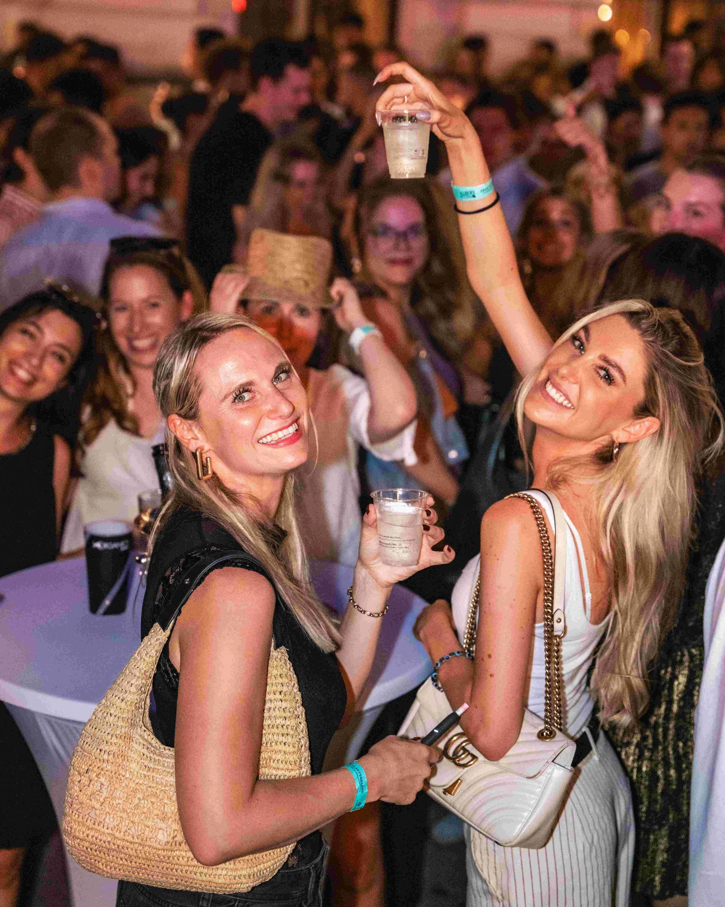 Two smiling women at a crowded social event, one holding a cup with a beverage, and the other with a white purse and striped outfit. The background shows many people in an indoor setting.