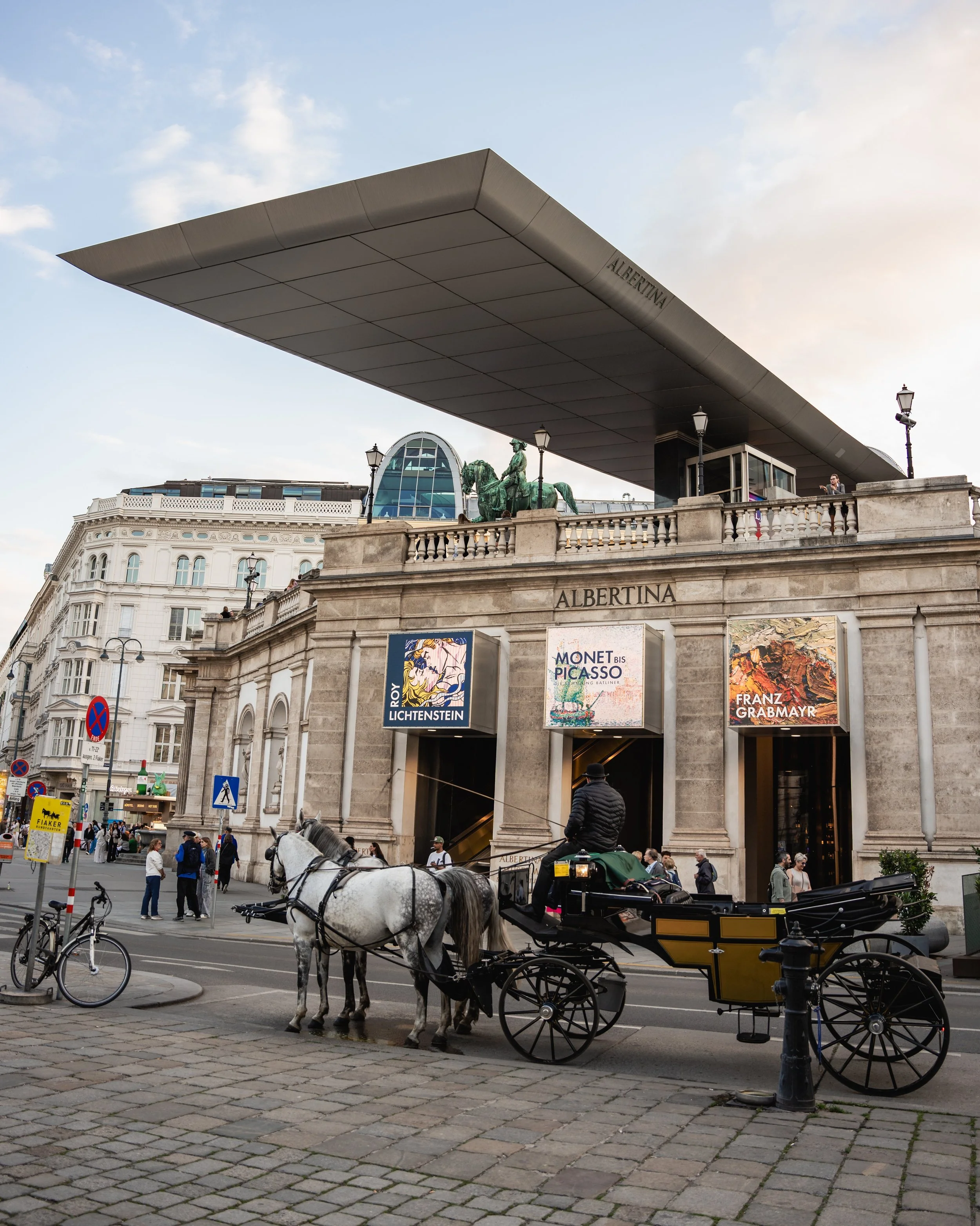 The image depicts the exterior of the Albertina Museum in Vienna, Austria. There is a large modern overhang at the entrance, with banners advertising exhibitions featuring Roy Lichtenstein, Monet and Picasso, and Franz Grabmayr. The facade includes c