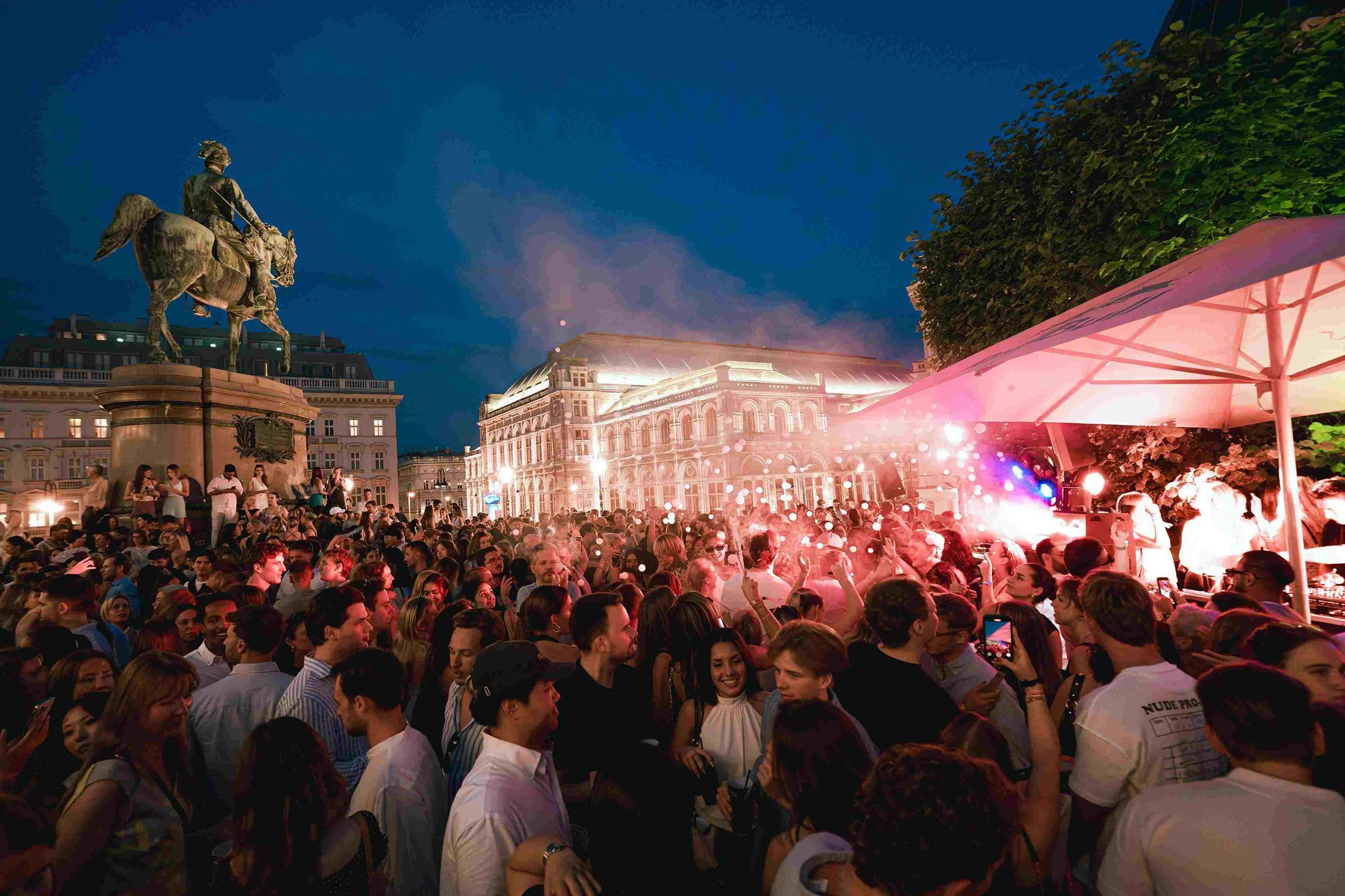 Crowd of people attending an outdoor evening concert or festival, with a statue of a man on horseback to the left and illuminated historic buildings in the background, a pink-lit performance area on the right.