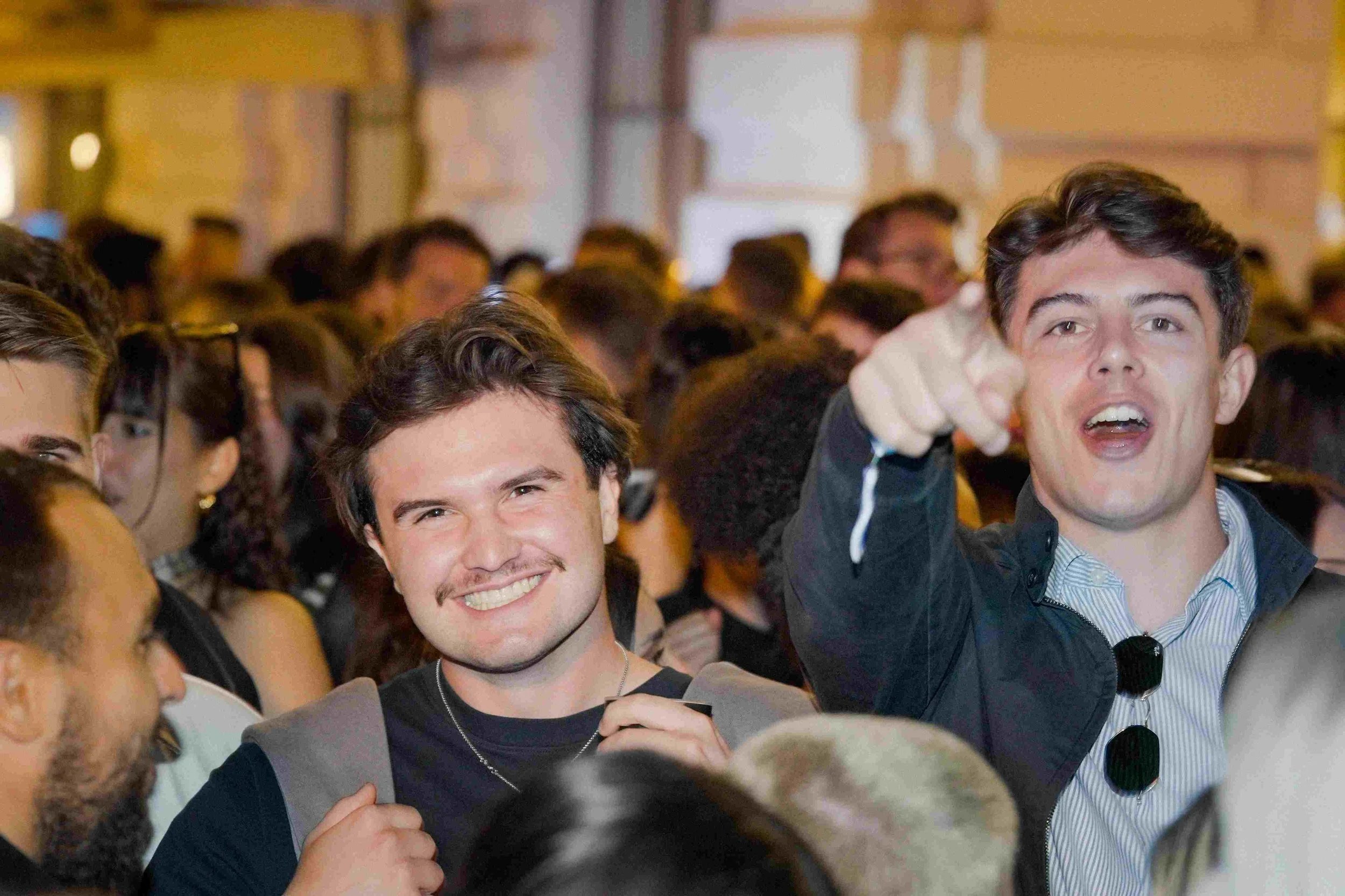People attending a crowded indoor event, smiling and engaging with each other, with a focus on two young men in the foreground who are posing and smiling at the camera.
