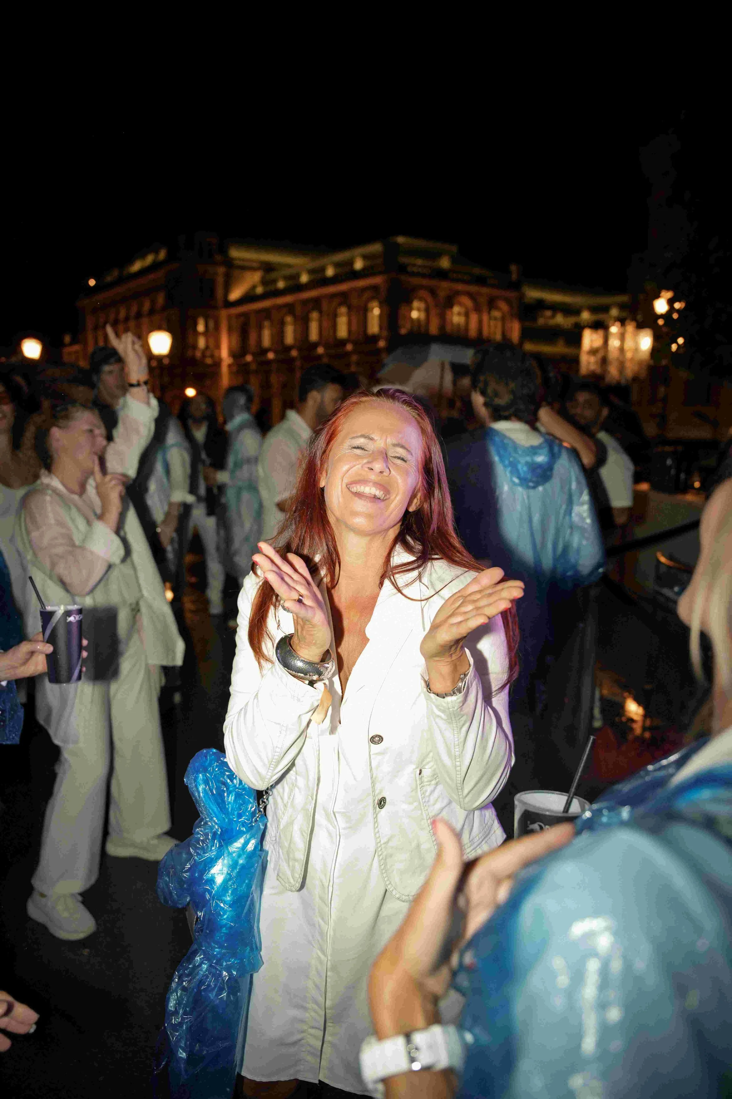 A woman with red hair and a white jacket is smiling with her eyes closed at night, surrounded by people in casual and rain gear, some holding drinks, outside a lit building.