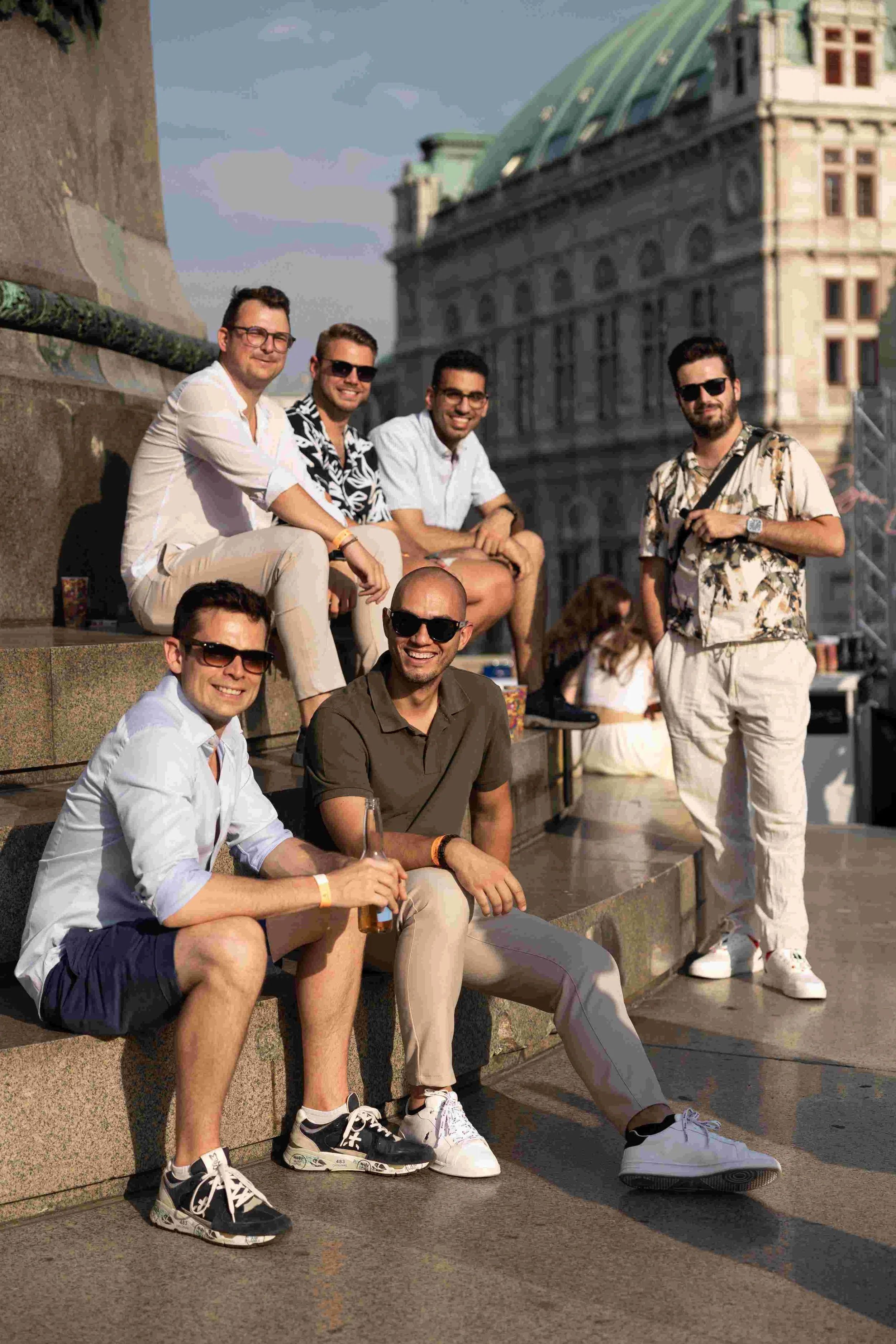 Group of seven young men sitting and standing on stone steps outdoors, with historic building in background, dressed casually in summer clothes, some wearing sunglasses, smiling and engaging with each other.