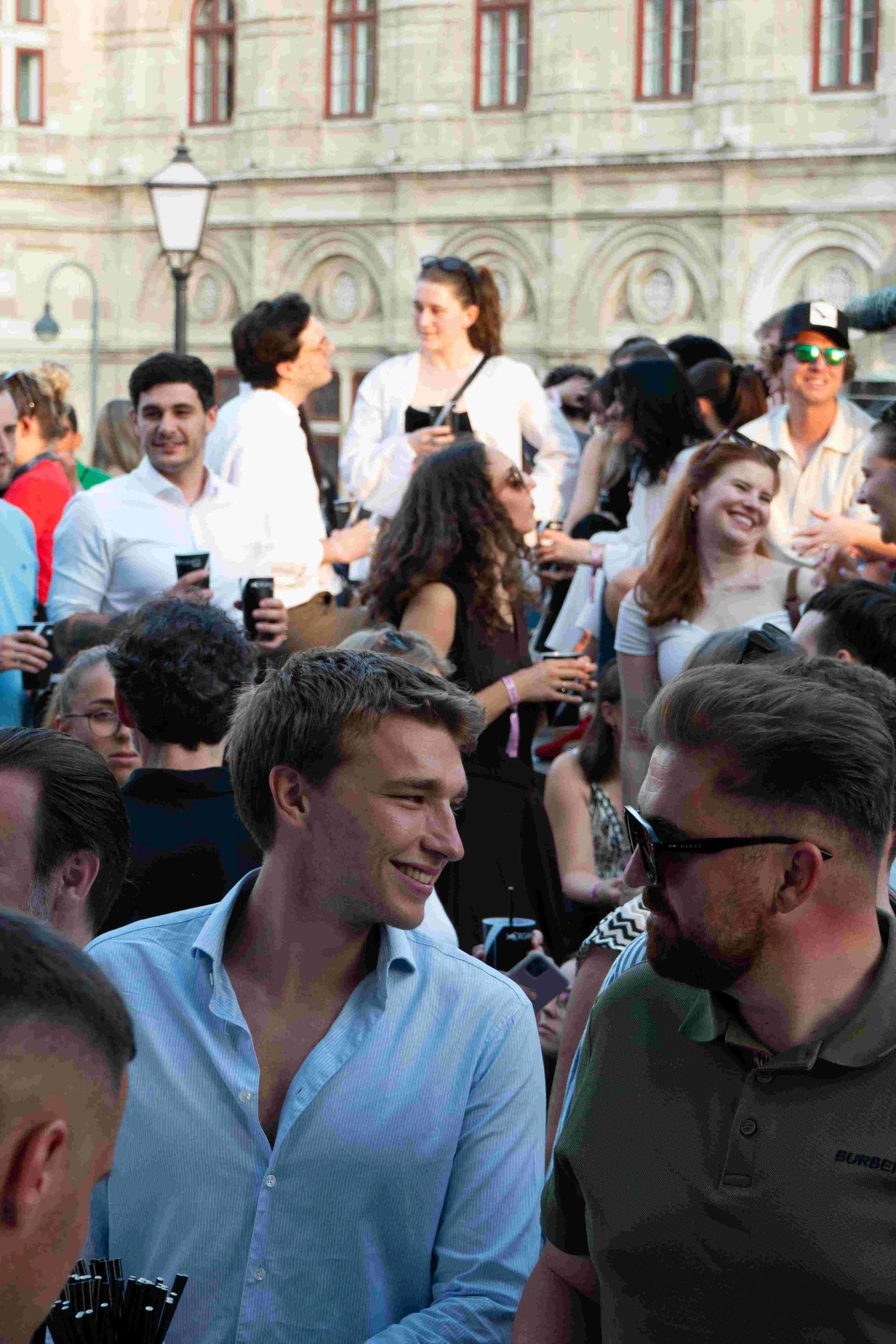 Group of people gathered outdoors in front of an old building, some smiling and holding drinks, engaged in conversation.