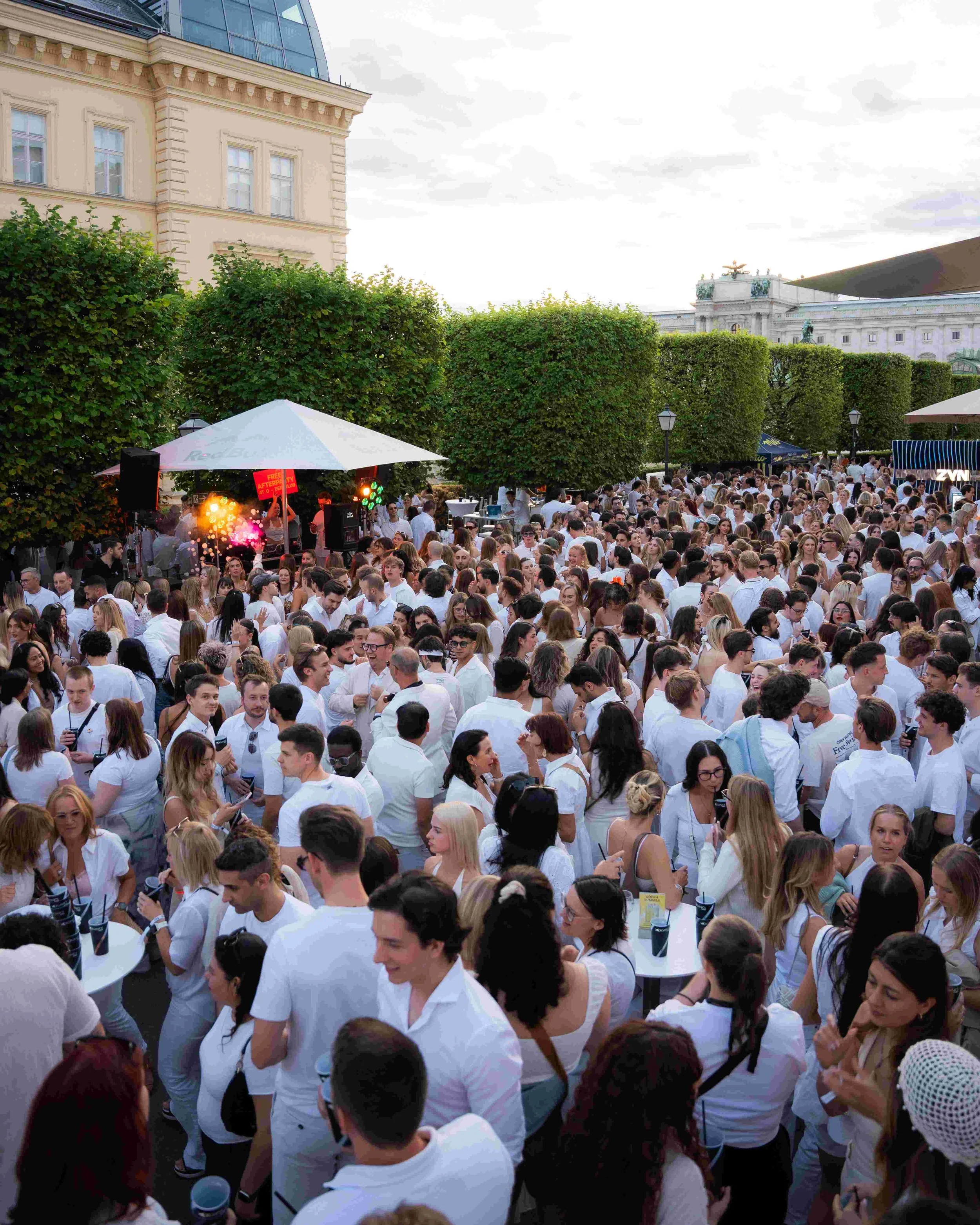Crowd of people gathered outdoors during the daytime, many wearing white, with some holding beverages and engaging in conversation. In the background, there are lush green trees and historic buildings, including one with ornate architectural details.