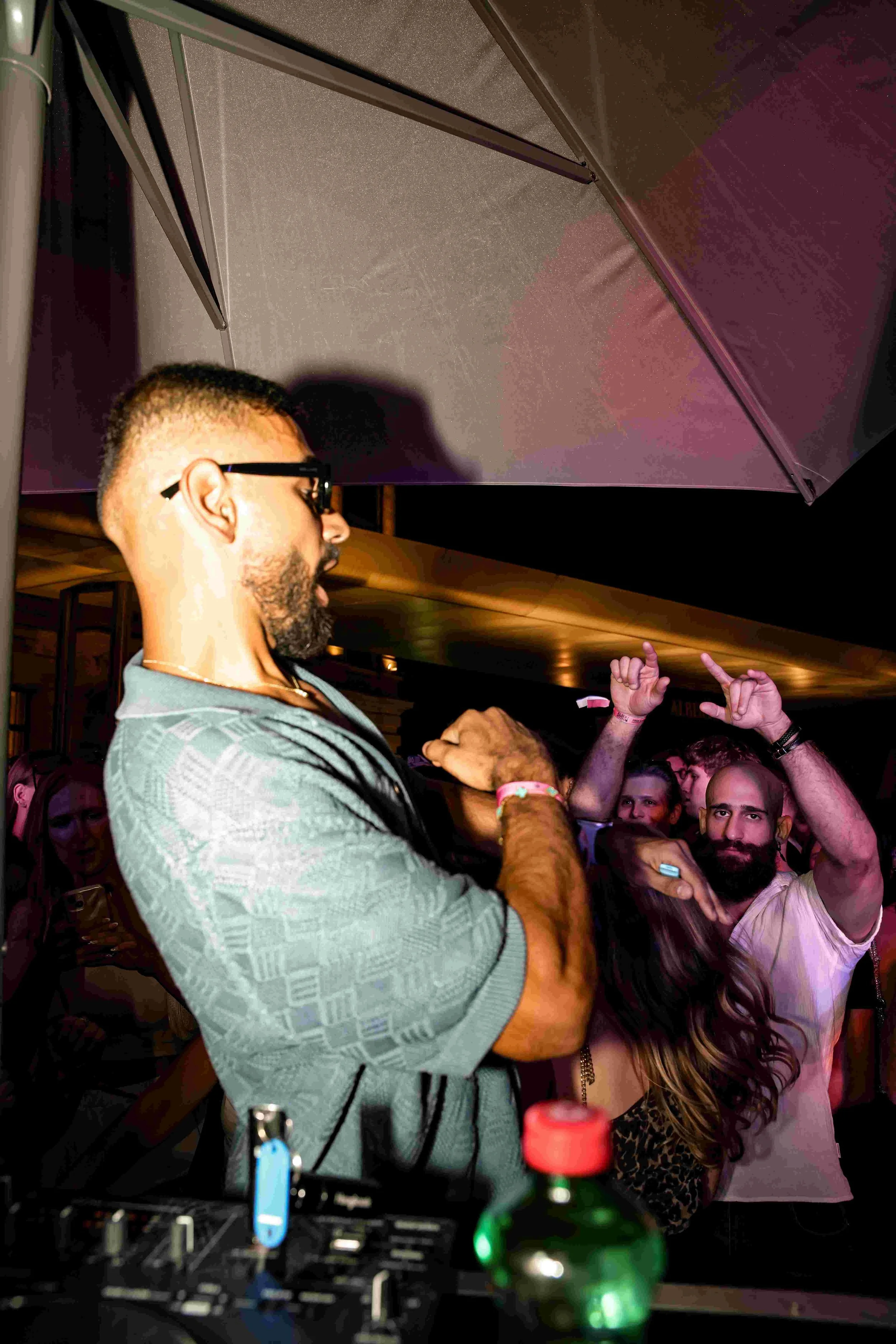 A man with glasses and a gray patterned shirt stands at a party or club with a DJ mixer in front of him and several people dancing and socializing in the background. Some people are making hand gestures and appear to be enjoying the music.