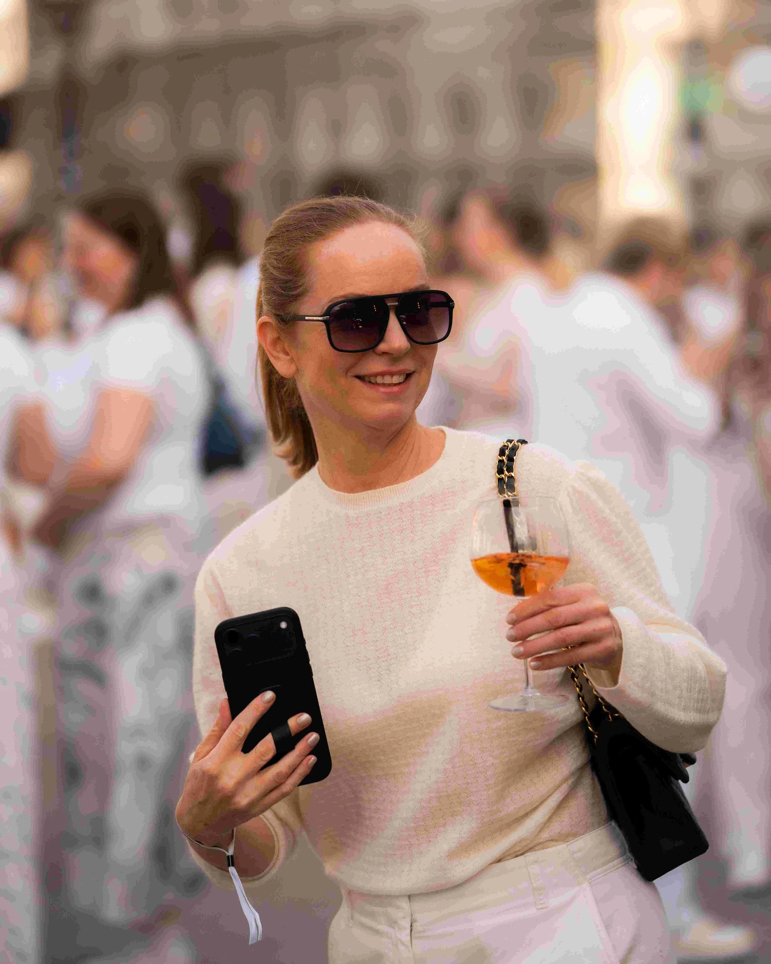A woman with sunglasses smiling while holding a glass of orange cocktail and taking a selfie with her smartphone in a crowded indoor setting.