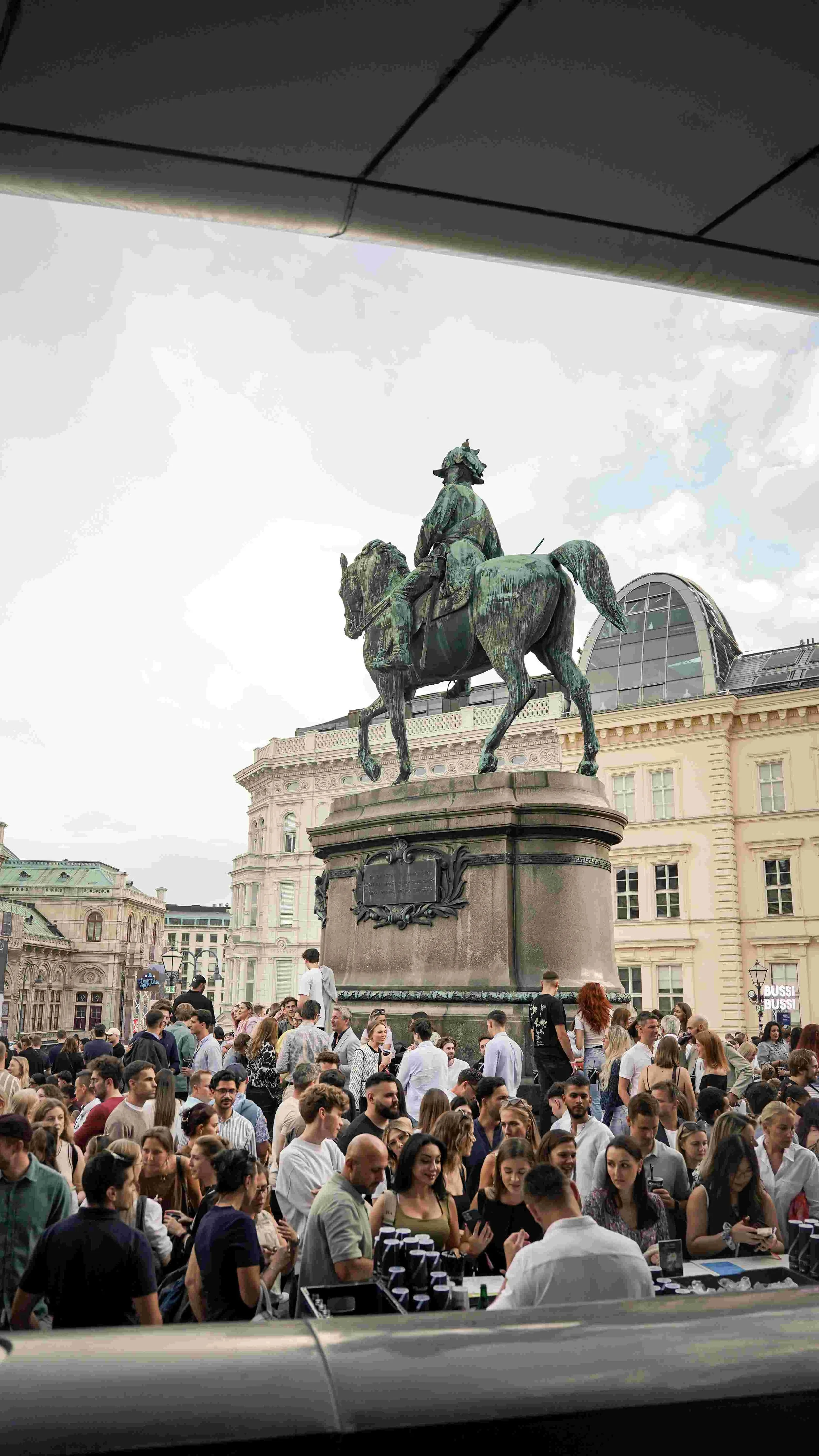 A crowded outdoor scene in front of an equestrian statue of a historical figure on a city square. Many people are gathered, some are talking, others are looking at their phones or buying items from a table. Surrounding buildings are historic and mode