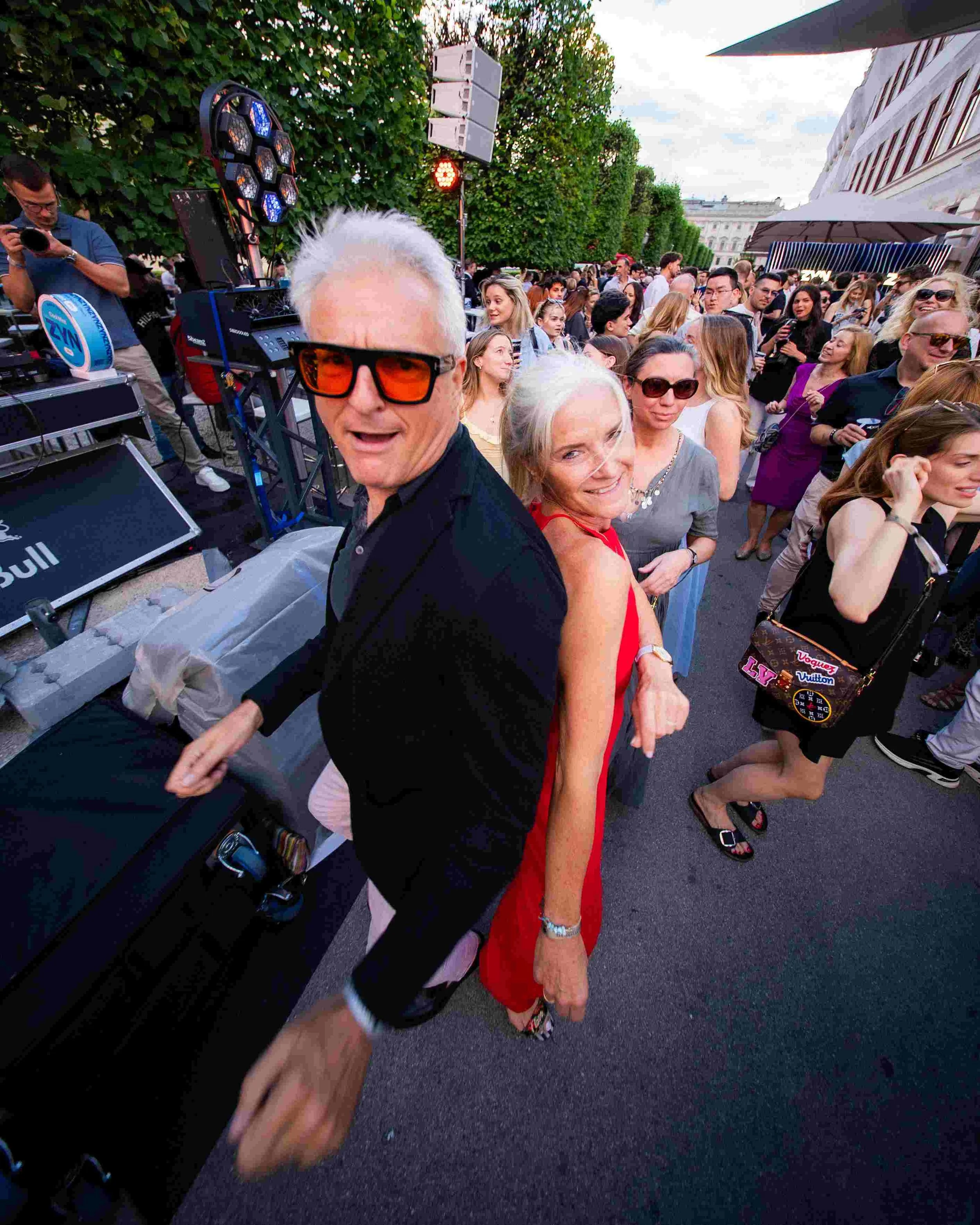 A lively outdoor event with a crowd of people, including an elderly woman in a red dress and sunglasses, a man with gray hair and sunglasses, and other attendees socializing. There is electronic equipment and lighting set up, and a man appears to be 