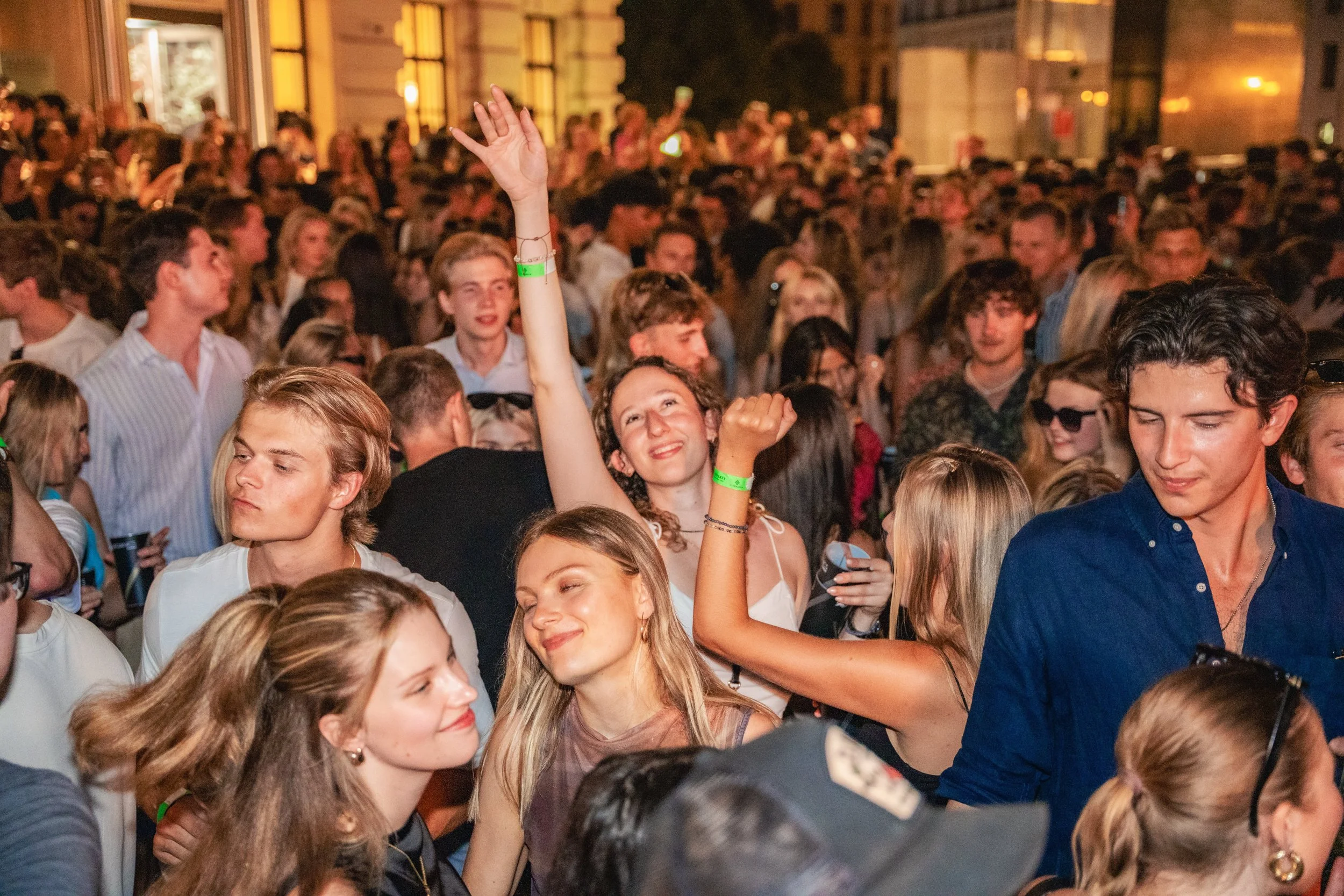 A crowded indoor concert or dance event with many young people, some wearing wristbands, enjoying the music and dancing.