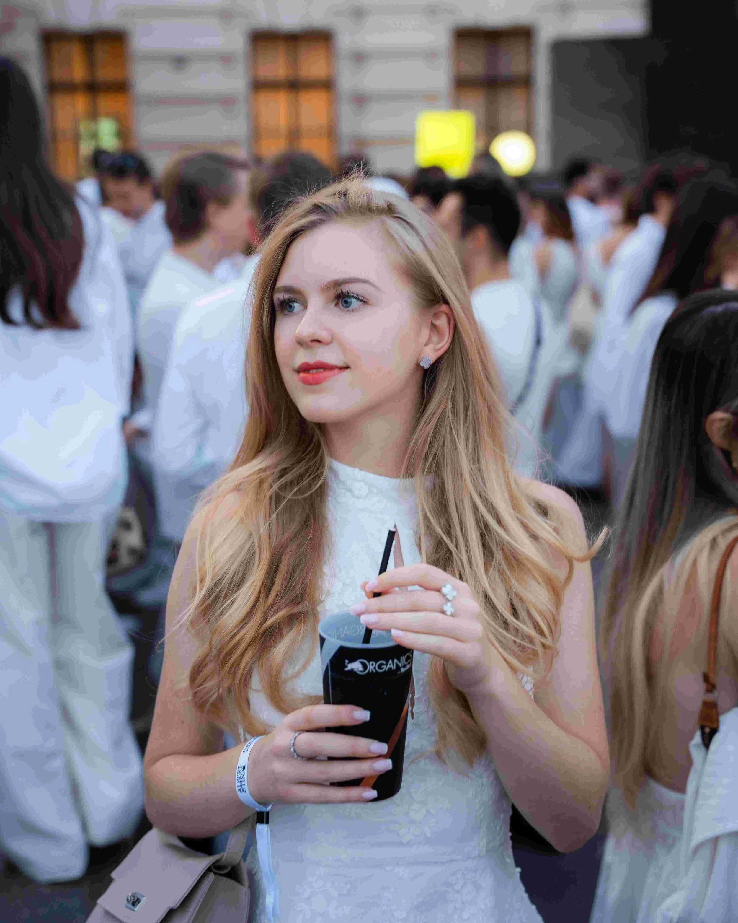 A young woman with long blonde hair, blue eyes, and light skin is holding a large cup of dark soda with a straw during an outdoor event with many people in white clothing in the background.