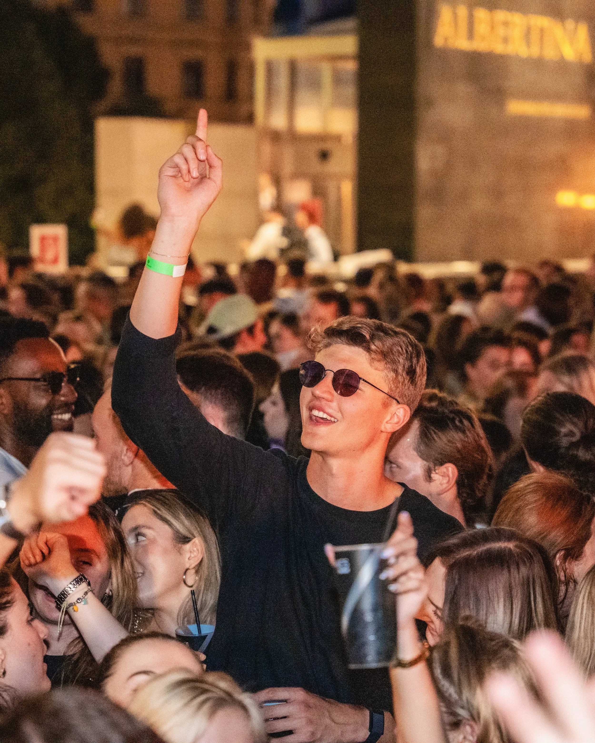 Crowd of people attending an event or concert, with a woman raising her hand, wearing sunglasses and a black top, smiling and surrounded by others. The background shows a sign with the word 'AUDITORIUM' and dim lighting.