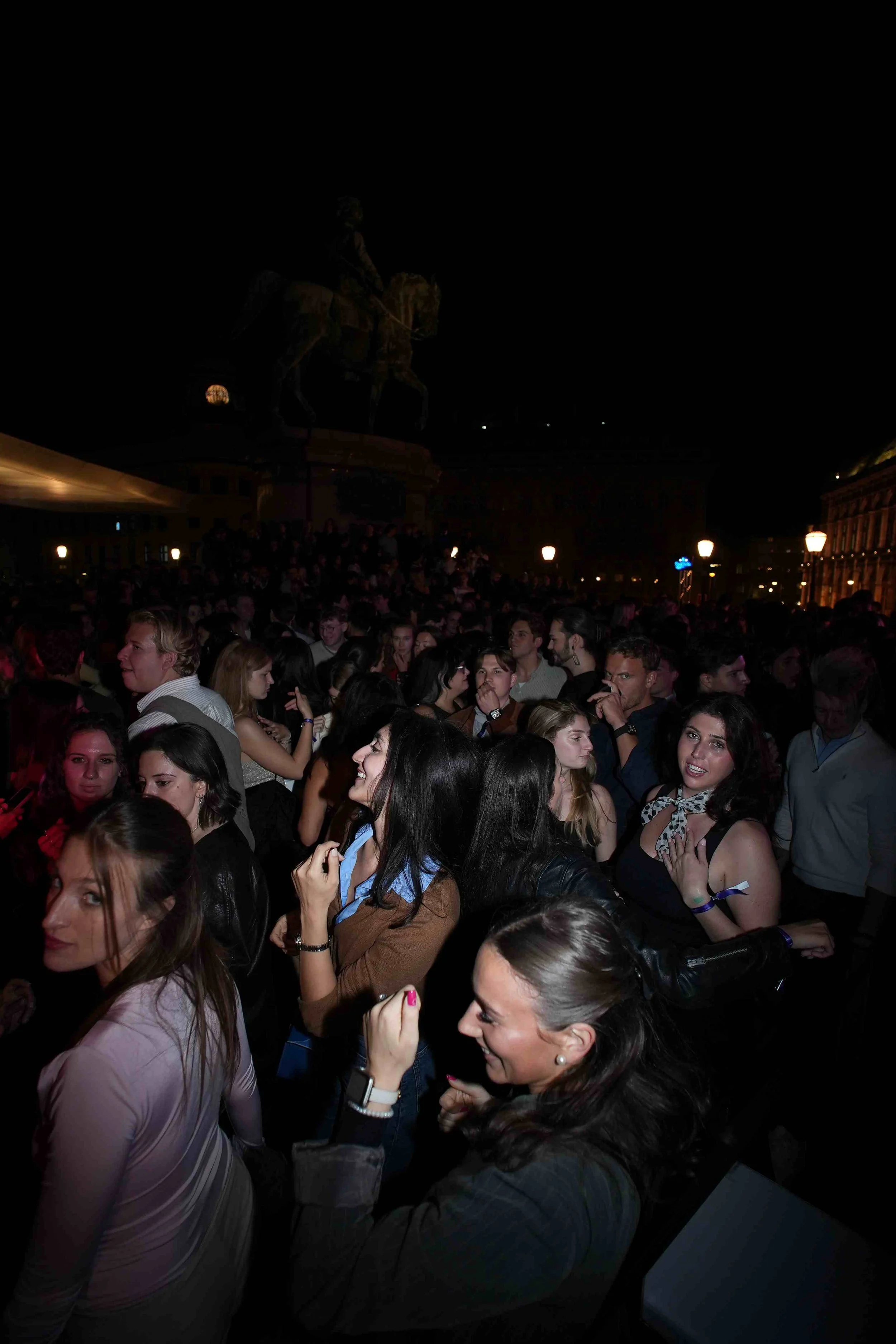A large crowd of people gathered outdoors at night, with a dark sky and some illuminated buildings and streetlights in the background. Some individuals are smiling and engaging with each other.