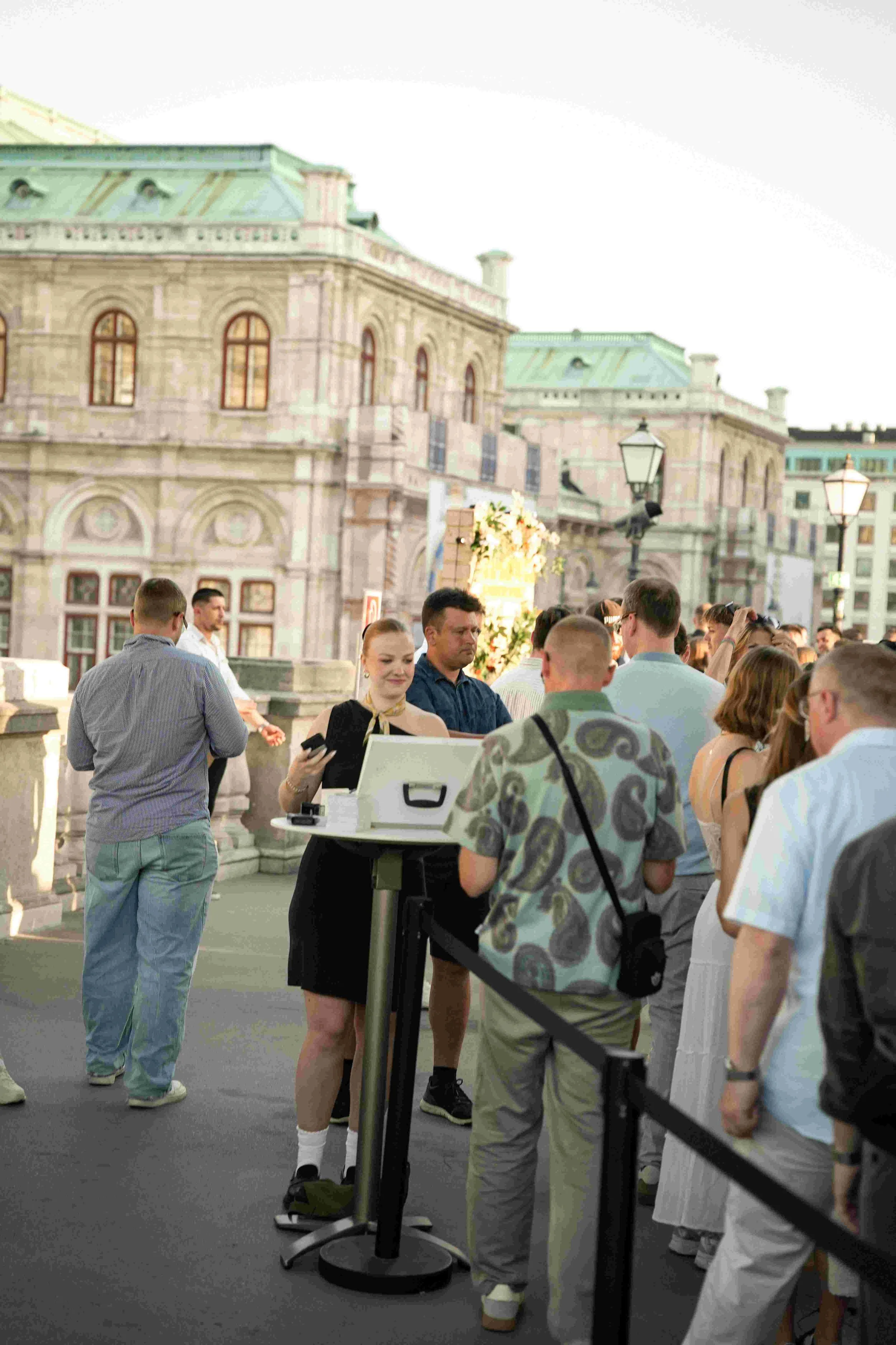 People gather outside a historic building with ornate architecture, which appears to be a palace or government building. Some individuals are standing in line or engaged in conversation. The scene takes place during daylight hours.