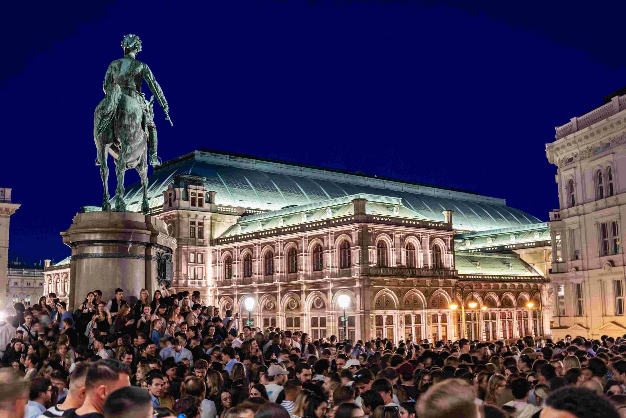 A large crowd of people gathered in an outdoor historical square at night, with a statue of a person on horseback in the foreground, historic buildings illuminated in the background, and a clear dark sky above.