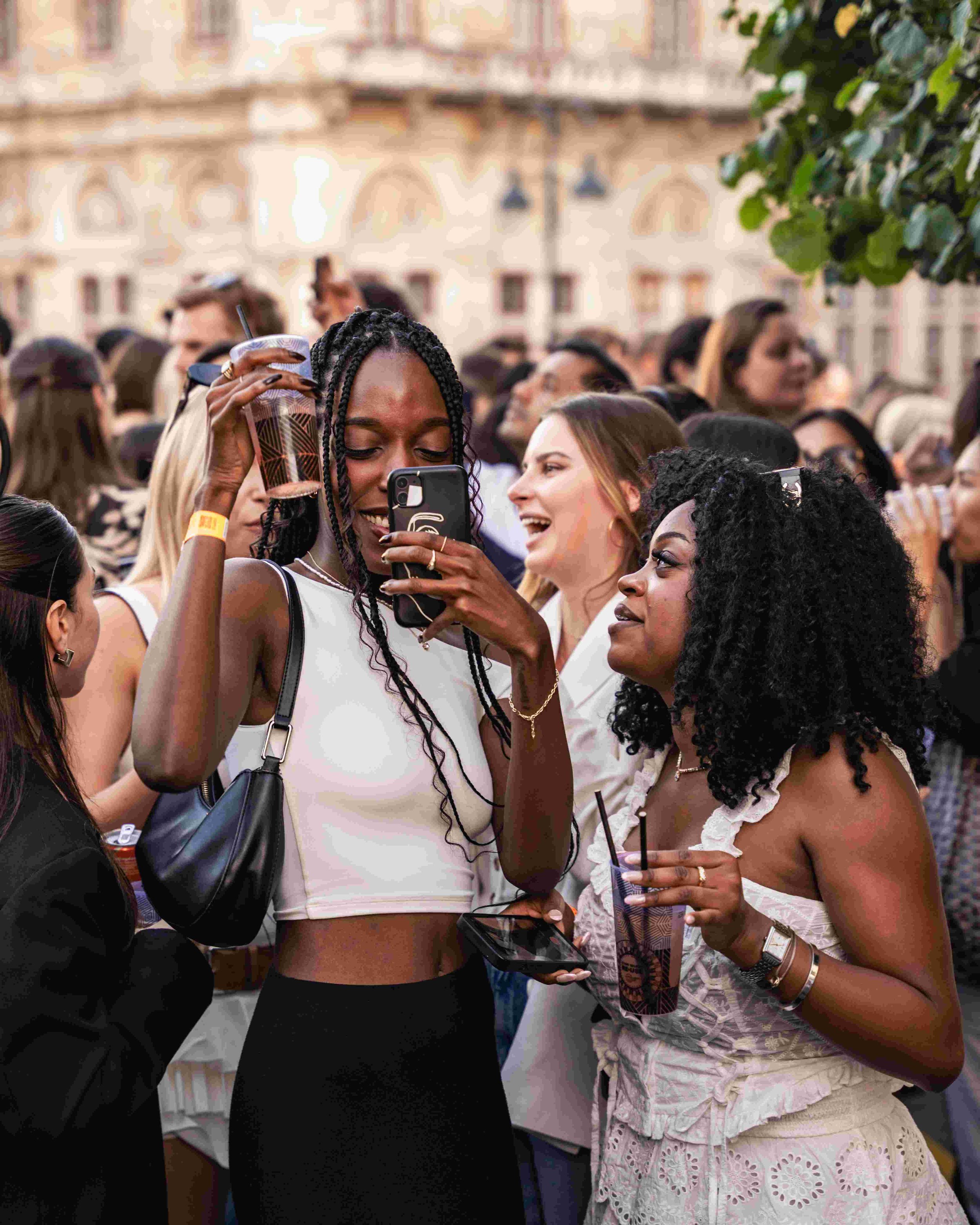 A diverse group of people gathered outdoors, smiling, talking, and taking photos with their smartphones, enjoying socializing in a city square with historic buildings in the background.