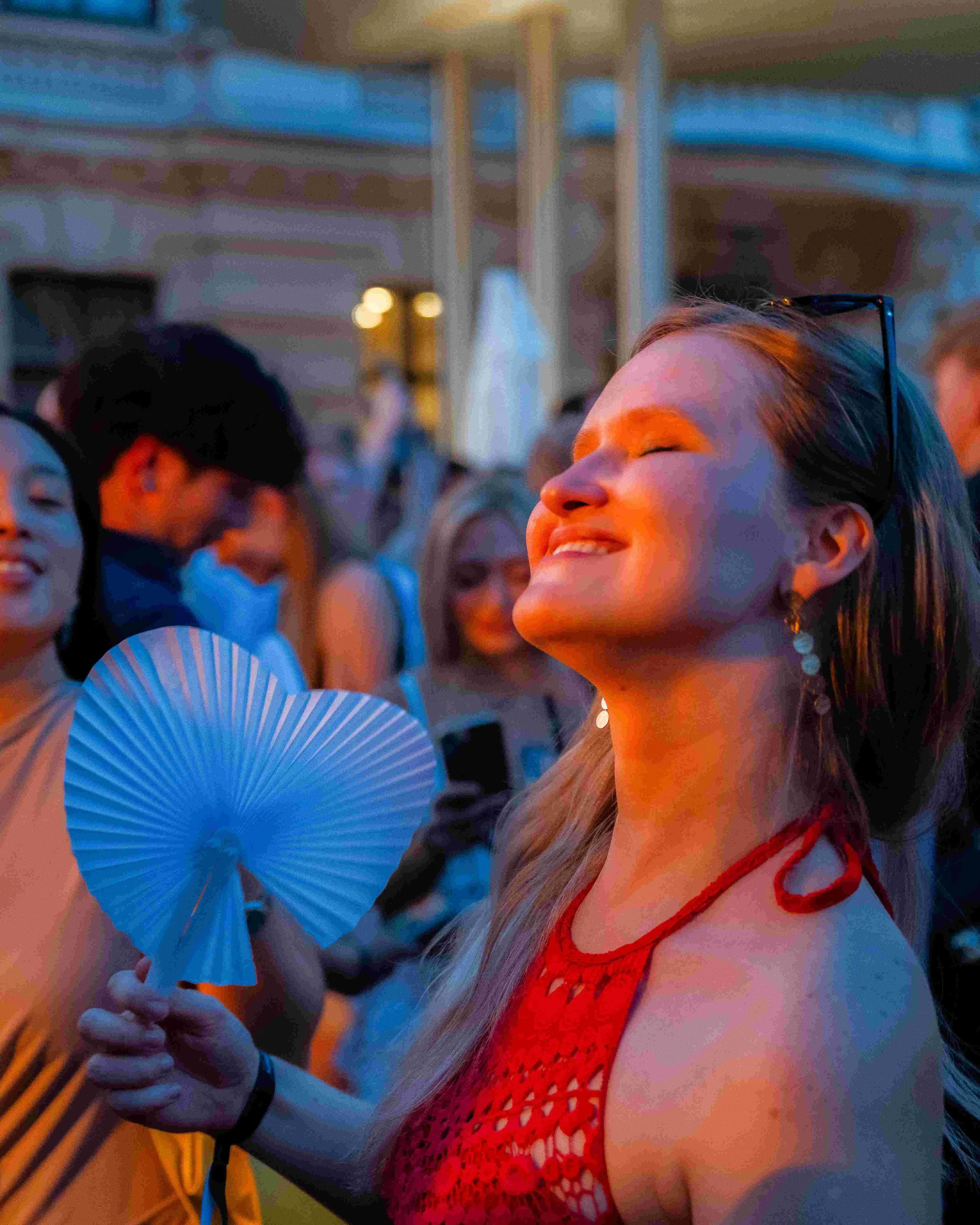 A young woman smiling with closed eyes, wearing glasses on her head, in a red lace dress, outdoors during twilight. Several other people are in the background, with colorful lighting.