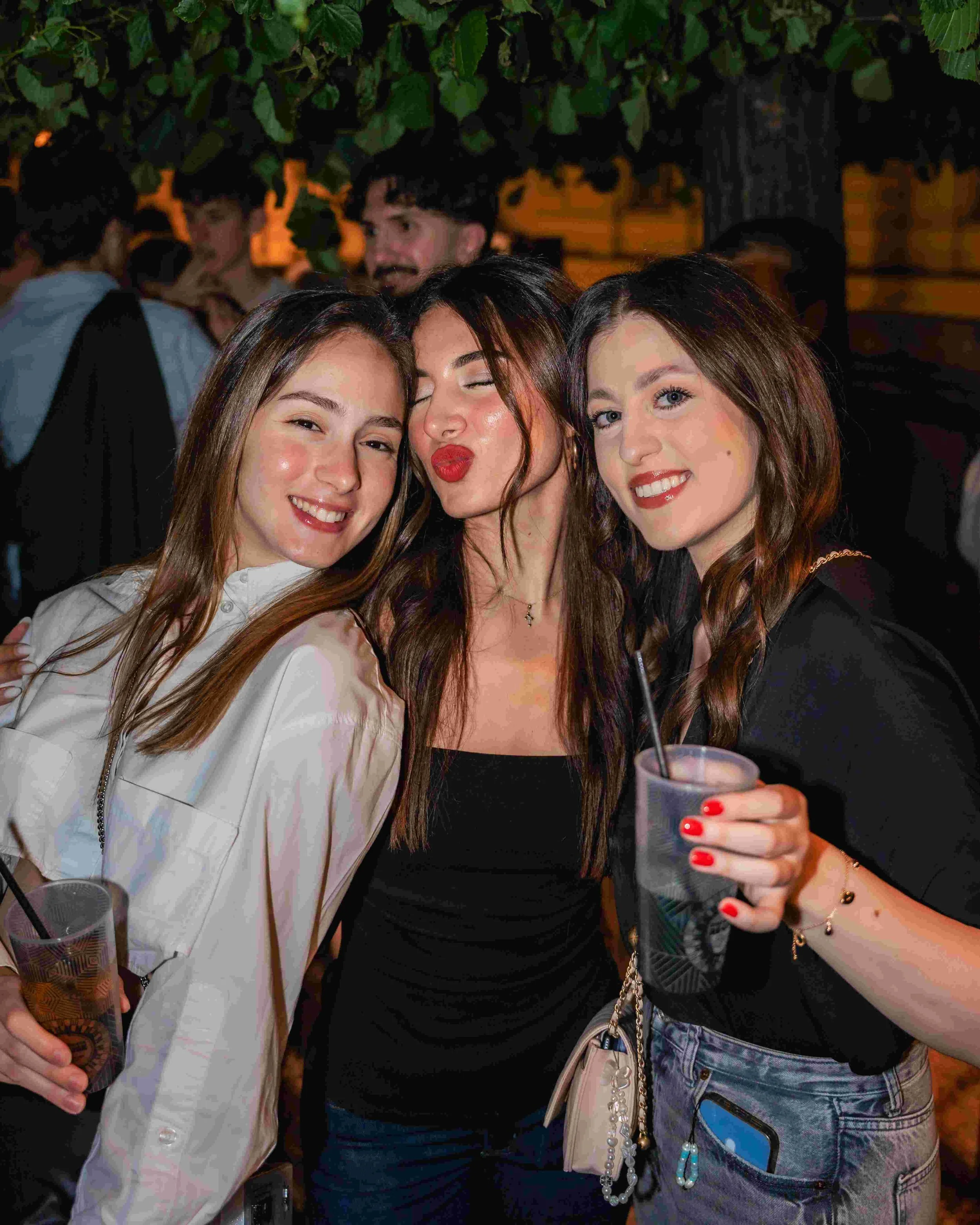 A group of young women are at an outdoor social gathering at night. Three women in the foreground are smiling and posing for the photo, with one woman in the middle making a playful face with her eyes closed and lips puckered. They are dressed casual