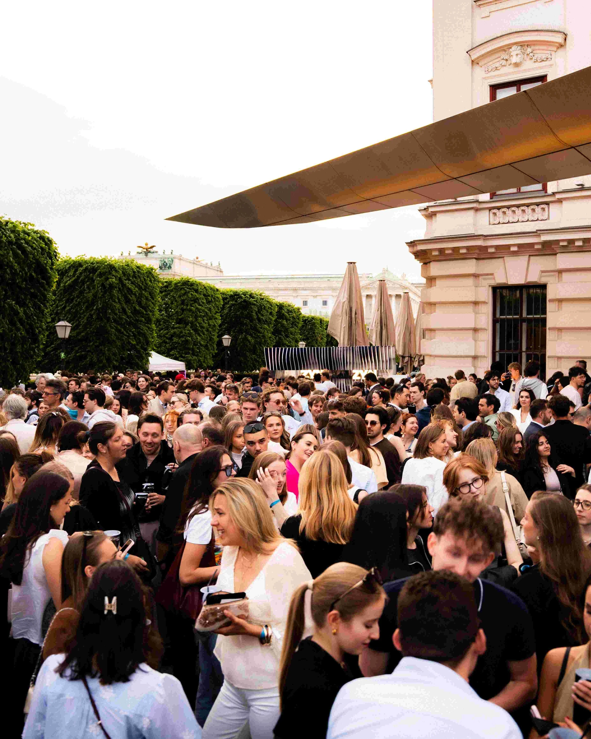 Crowd of people gathered outdoors near historic buildings with lush green trees and umbrellas, some people holding drinks and using mobile phones.