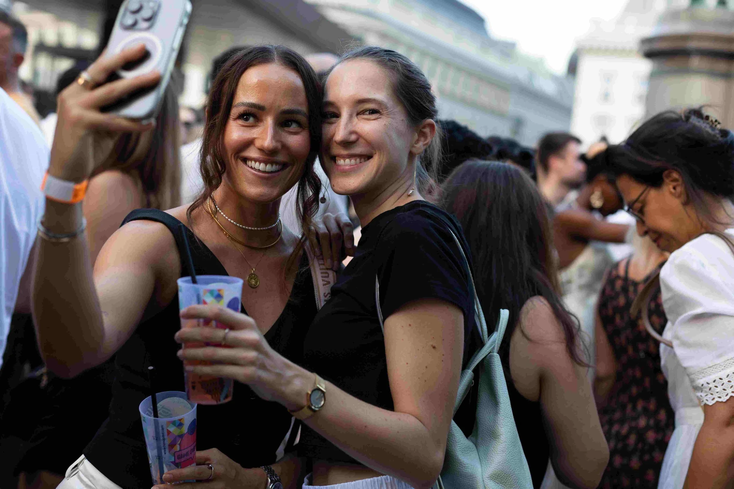 Two women smiling and taking a selfie at an outdoor event, surrounded by a crowd of people.