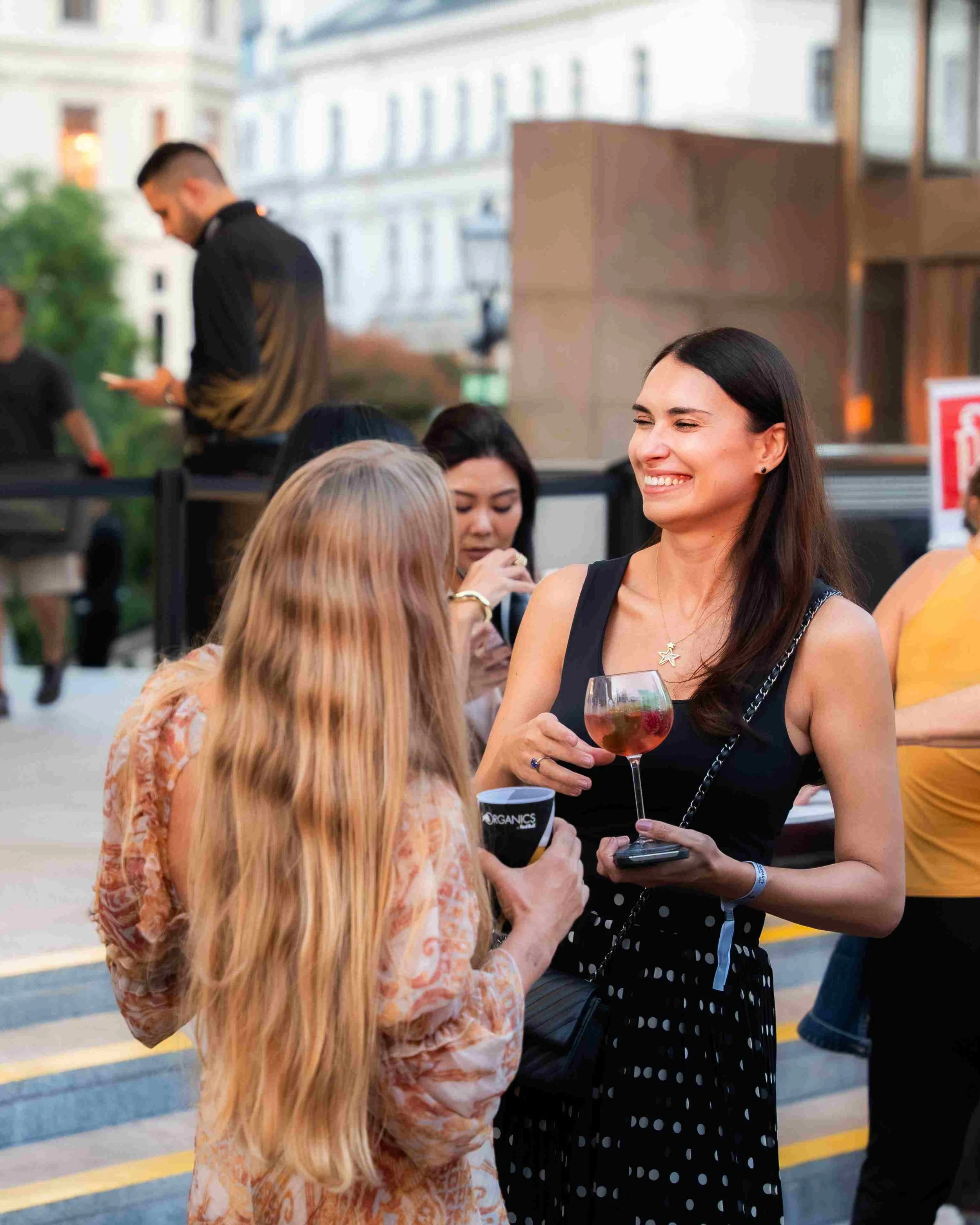 Two women are talking and smiling outdoors. One woman is holding a drink and the other is holding a cup. A man in the background is looking at his phone, standing near a building. It's evening or late afternoon.