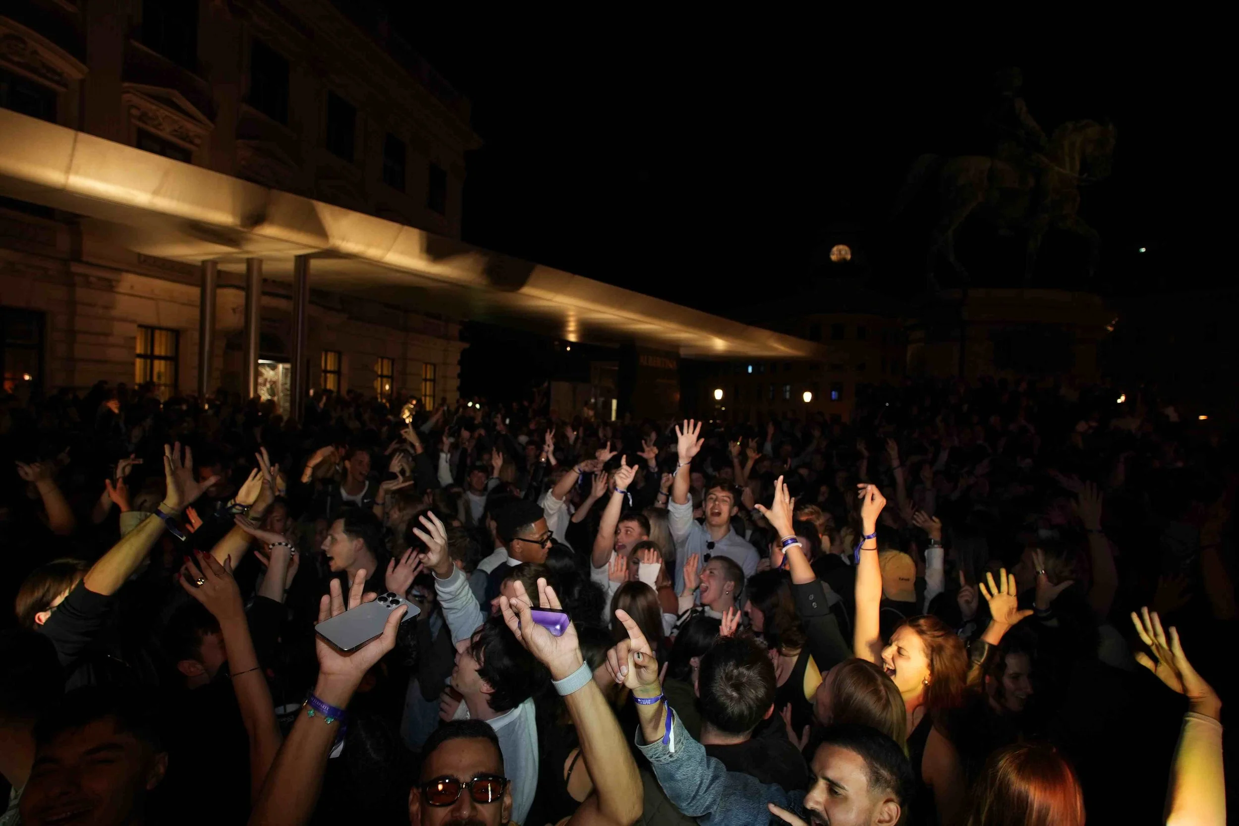 Crowd of people at a concert or event with some raising their hands, in a dark venue with a building facade and a large statue in the background.