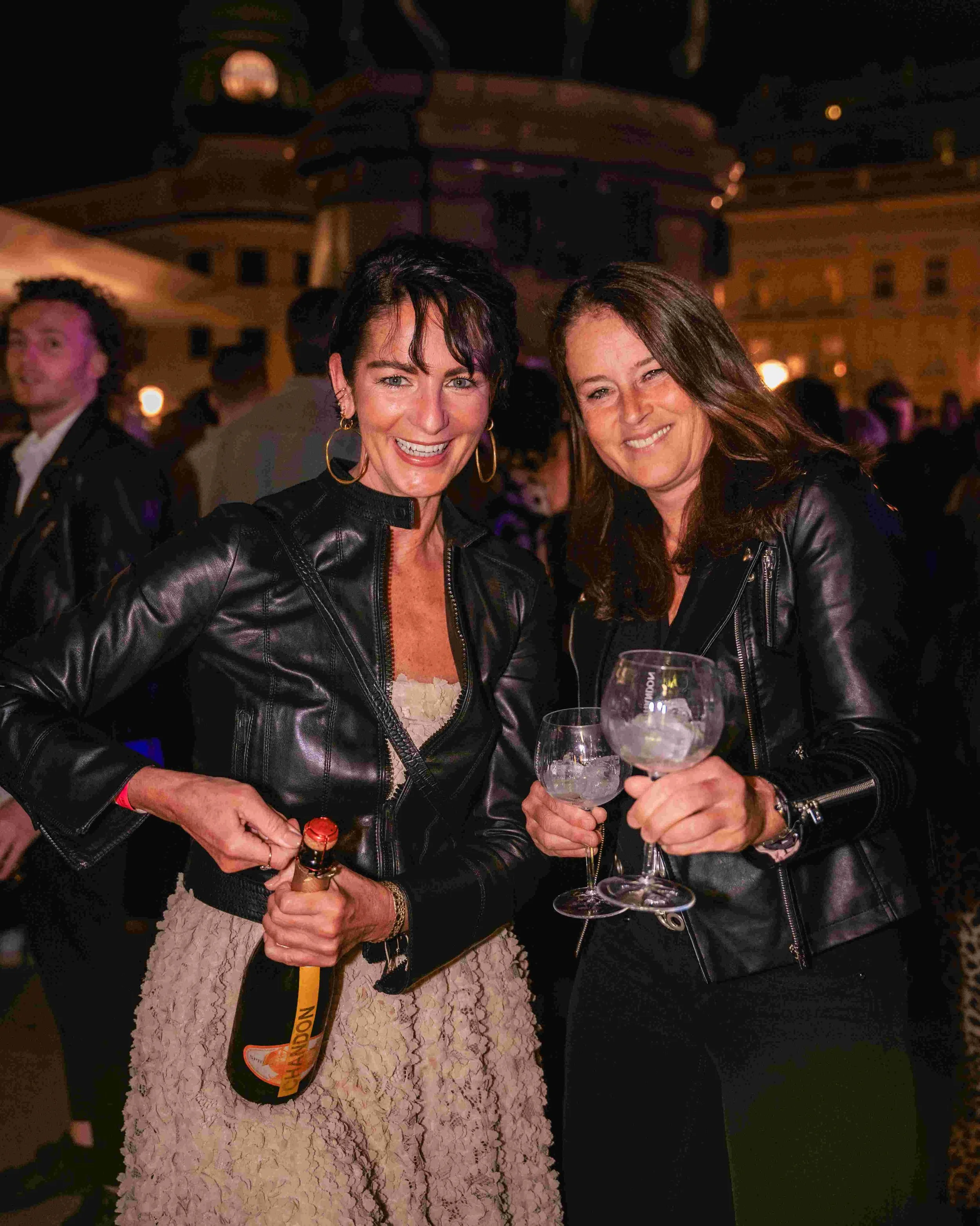 Two women at an outdoor event at night, smiling and holding wine glasses; one woman is wearing a leather jacket and the other a lace dress.