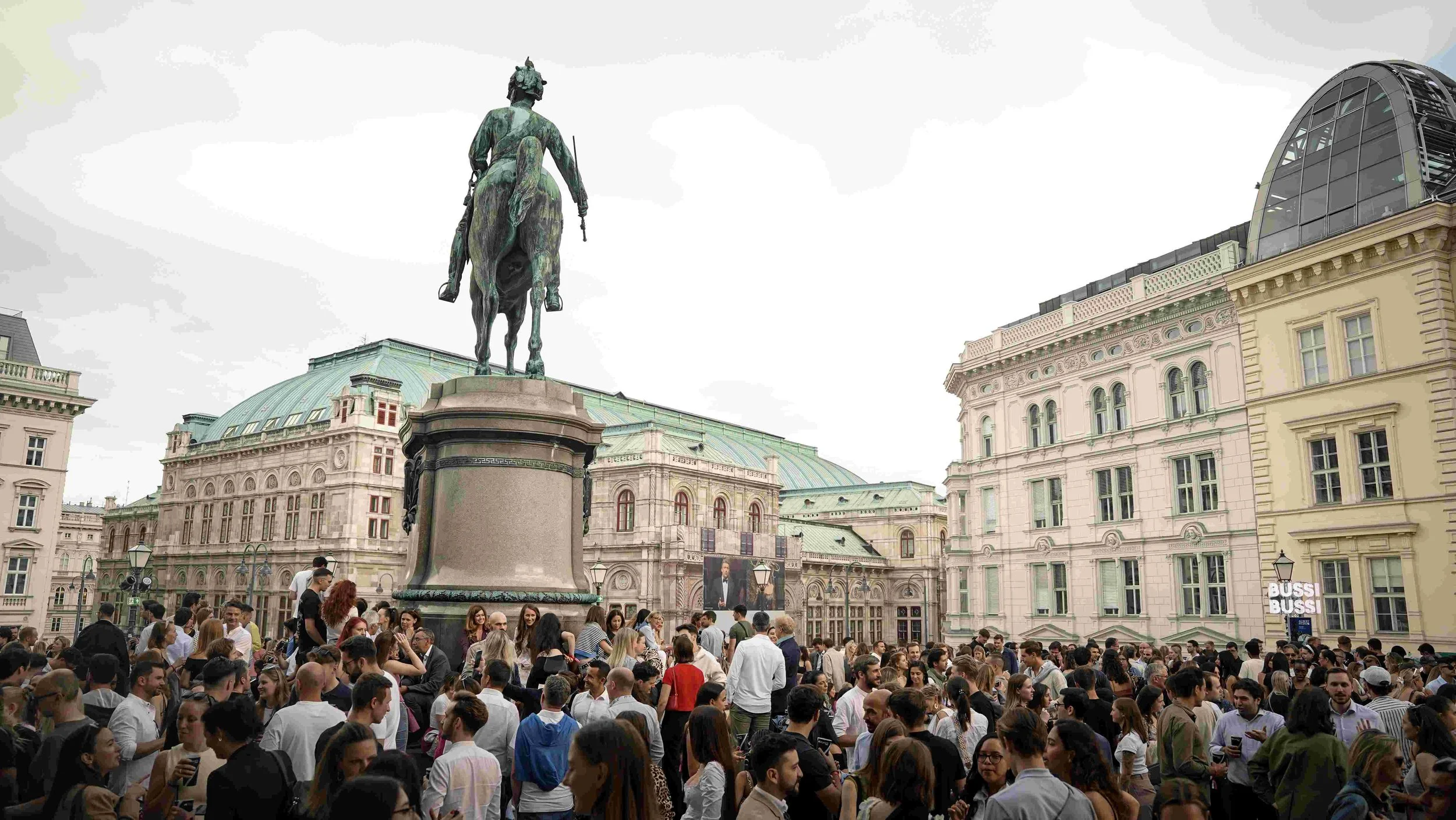 Large crowd gathered in a city square with historical buildings and an equestrian statue in the background.