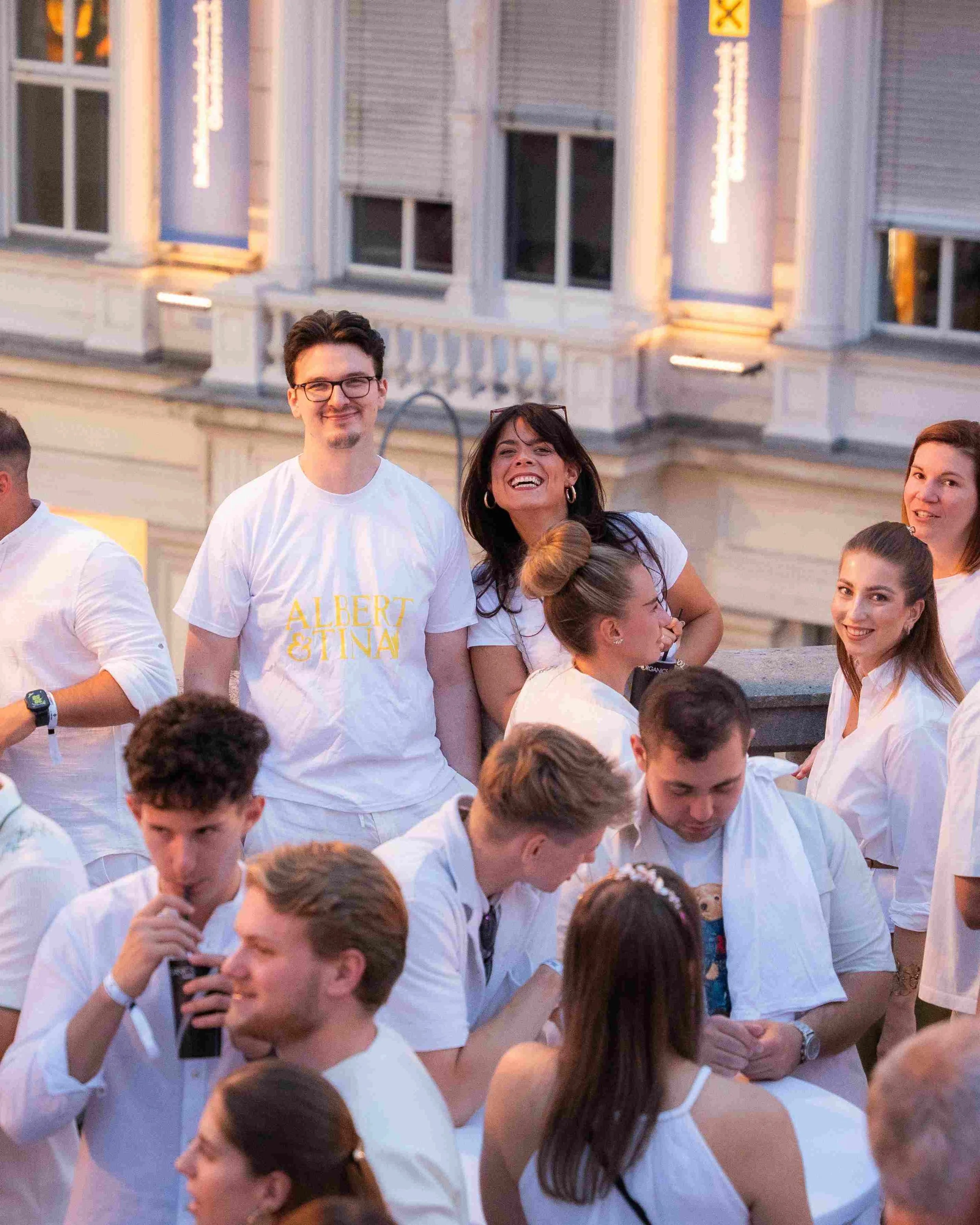 A group of people dressed in white shirts participating in an outdoor event or gathering in the evening, standing on a city street with a building in the background.