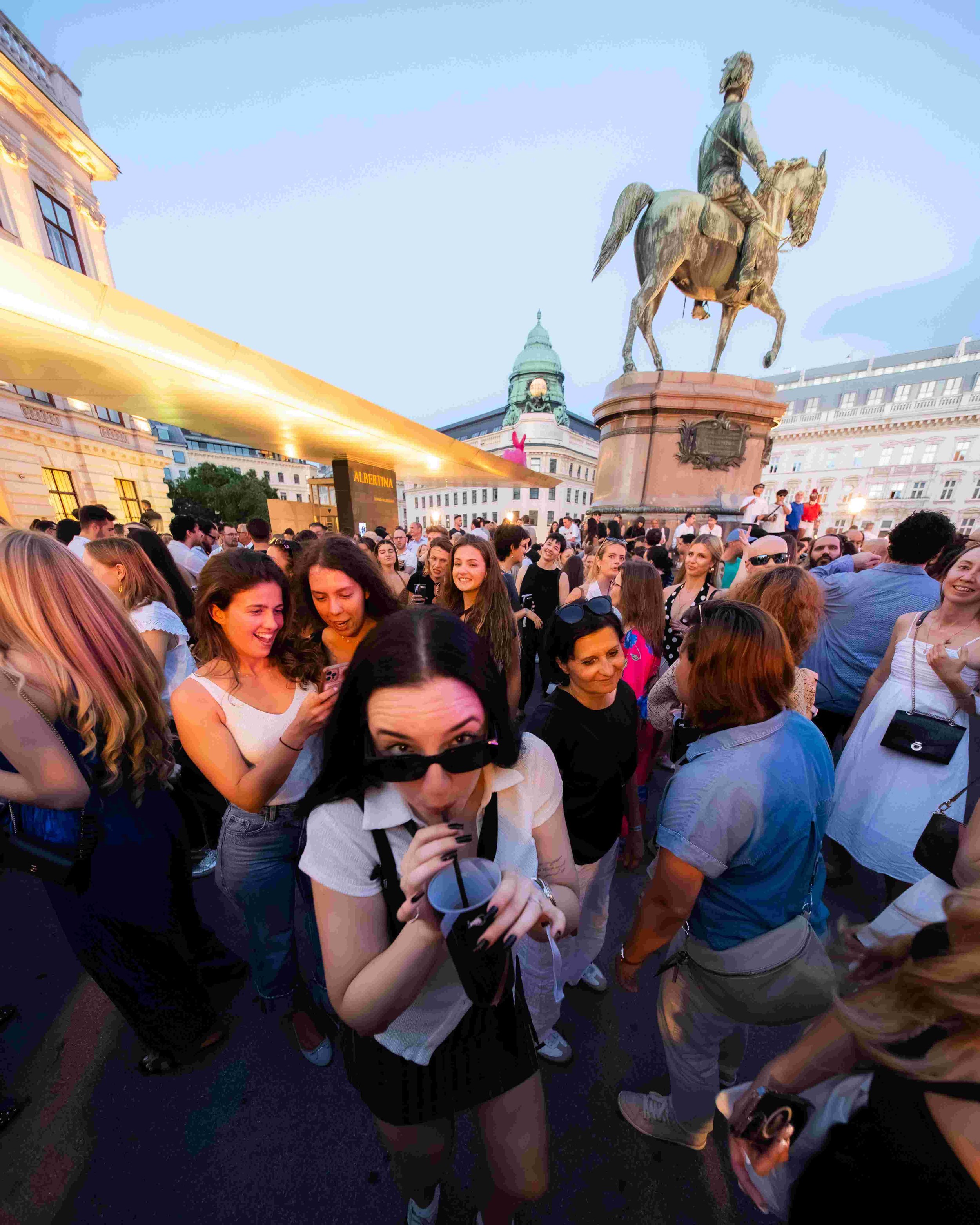 A crowd of people gathered outdoors in front of a large equestrian statue. The sky is clear and the scene is illuminated by evening light.