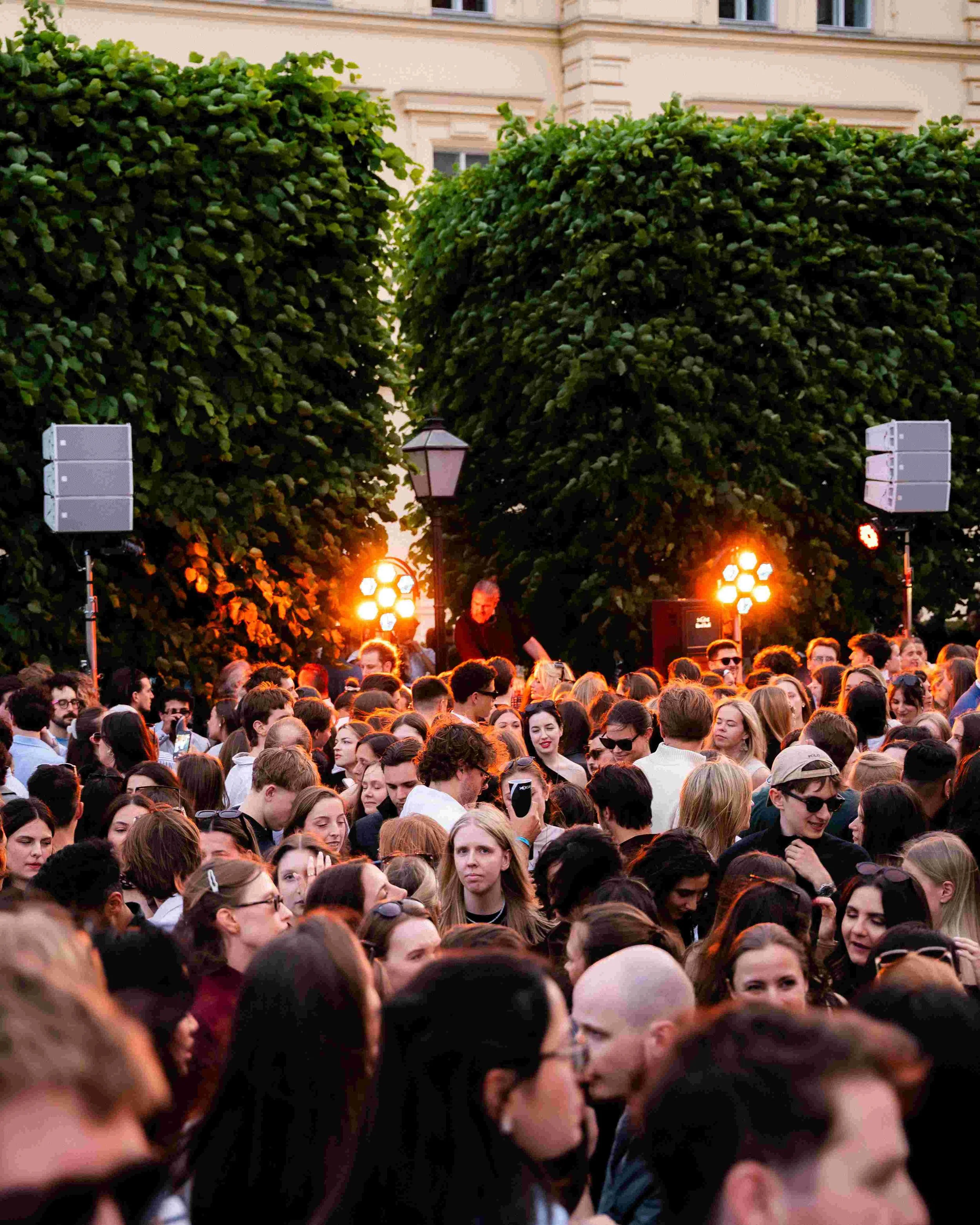 A large crowd of diverse people gathered outdoors during sunset, with two large speakers and stage lighting in the background, and a building with windows and greenery.