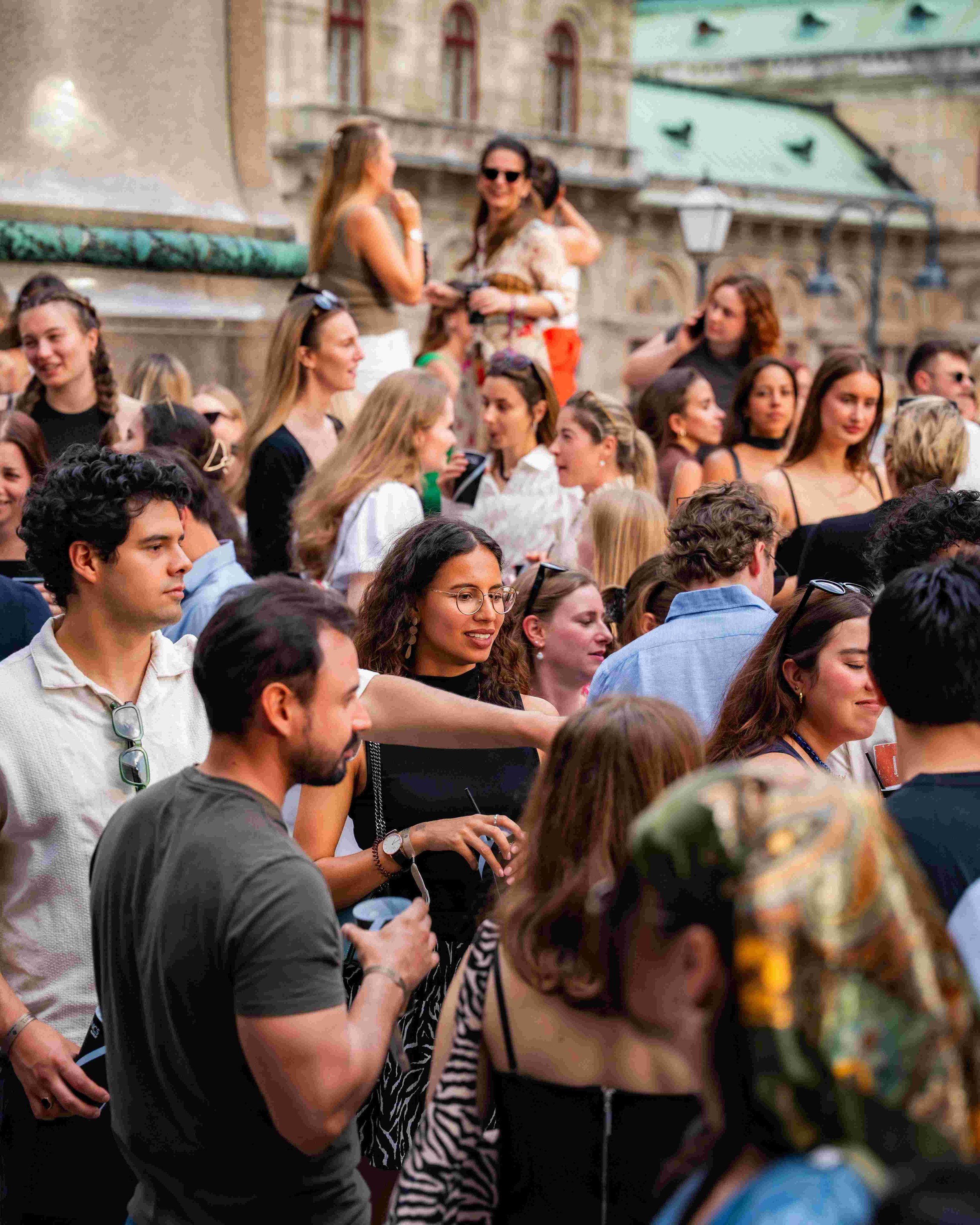 A large crowd of people outdoors, some talking and others looking on. Two women are standing on a ledge in the background, smiling and talking to each other, dressed casually, with an older building with arched windows behind them.