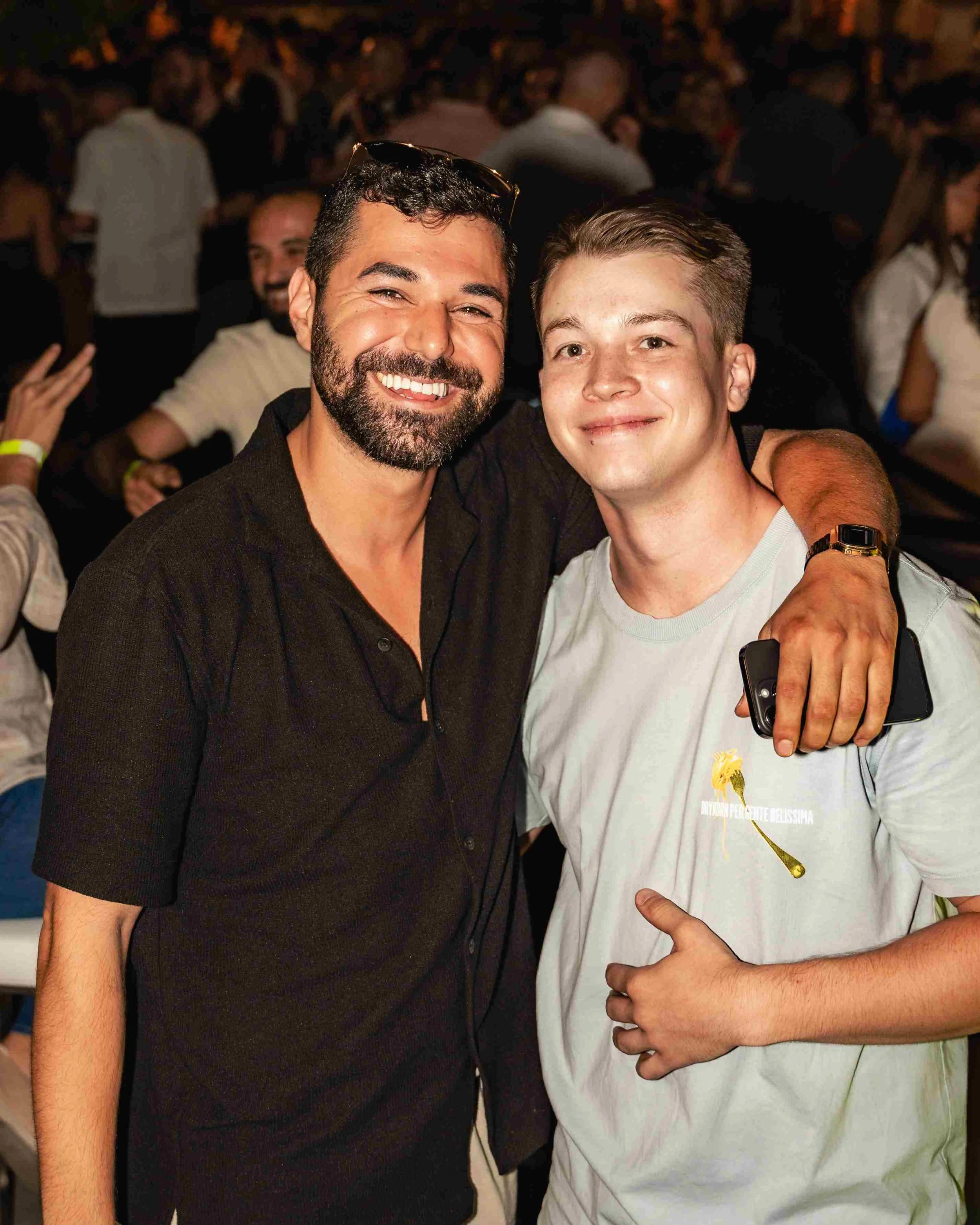 Two men smiling, one with a beard and sunglasses on his head, and the other with short hair, in a crowded setting with many people in the background, all appearing to enjoy a social event or gathering.
