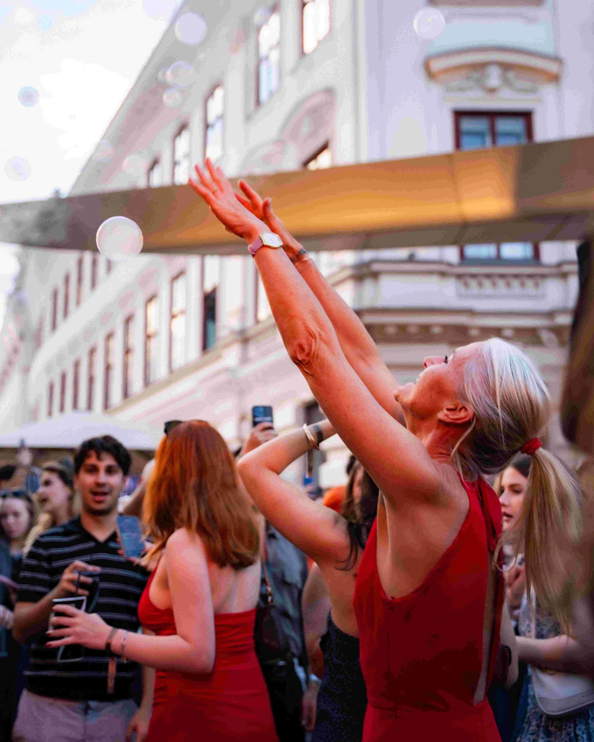 A crowd of people outside participating in a celebration or parade. Two women are dancing, one with white hair and the other with reddish hair, both wearing red dresses. The crowd looks joyful, some holding drinks, and one person is taking a photo. T