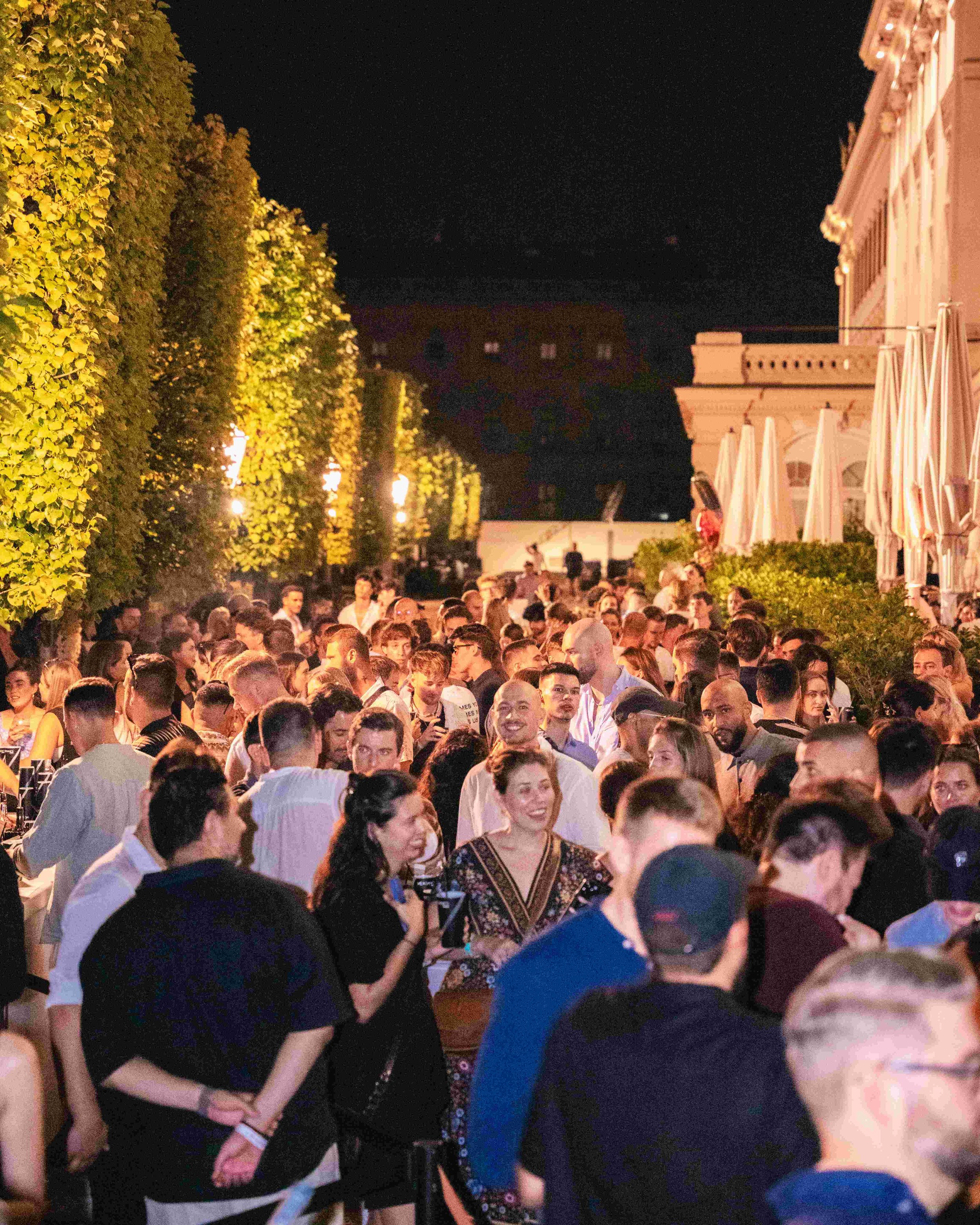 Crowded outdoor event at night with many people socializing. Green hedge on the left, white umbrellas on the right, building with decorative architecture in the background, and dark sky overhead.