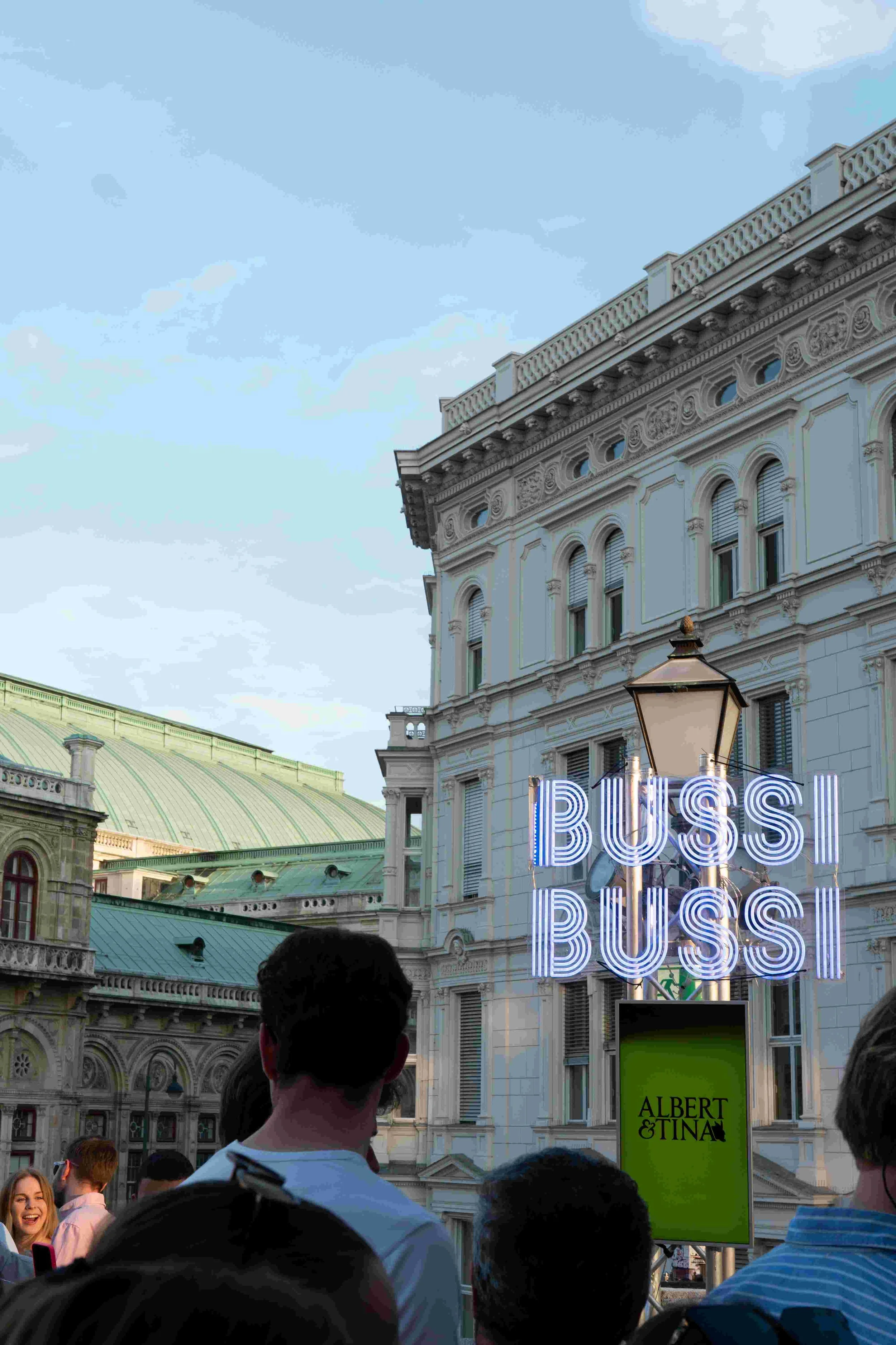 Crowd of people gathered in front of a historic building with detailed architecture and a green-domed roof, while neon signs display the word 'BUSS' and a green sign reads 'ALBERT & TINA'.
