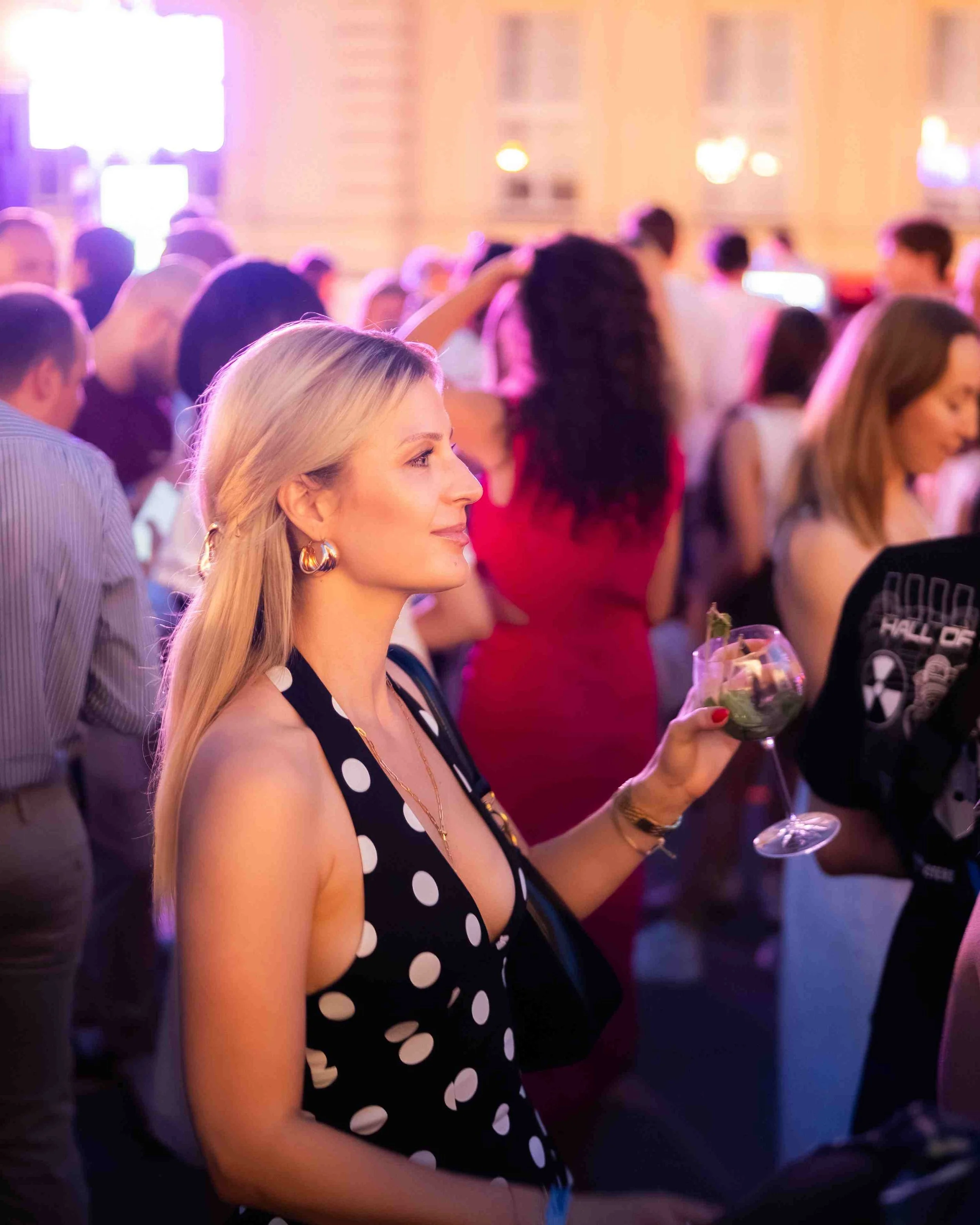 A woman with blonde hair and large earrings wearing a black dress with white polka dots, holding a glass of wine at an event with multiple people in the background.