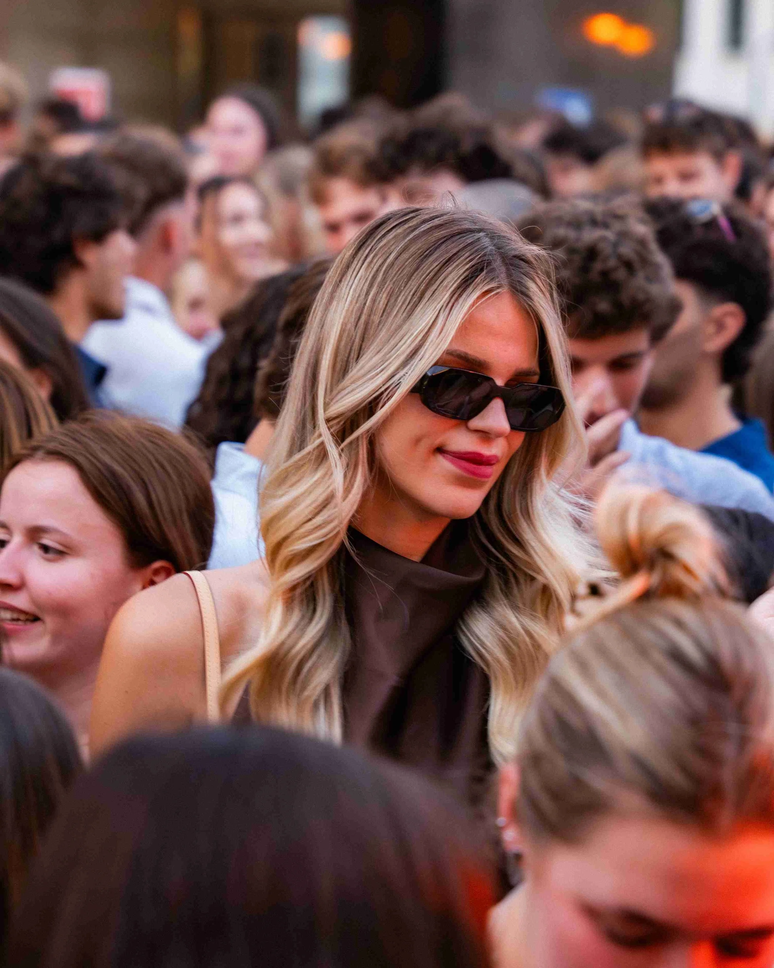 Blurry photo of a large crowd of people at an outdoor event, with a woman wearing sunglasses and blonde hair in the foreground.