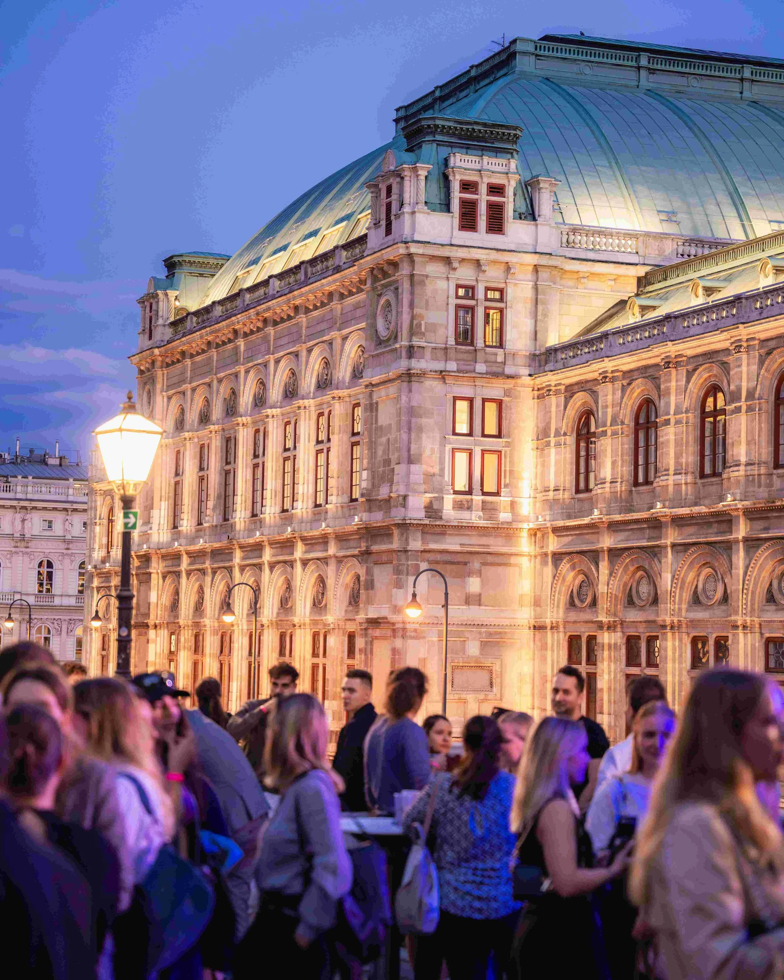 A large crowd of people gathered outdoors near a historic building with a domed roof at dusk, illuminated by streetlights and soft evening light.