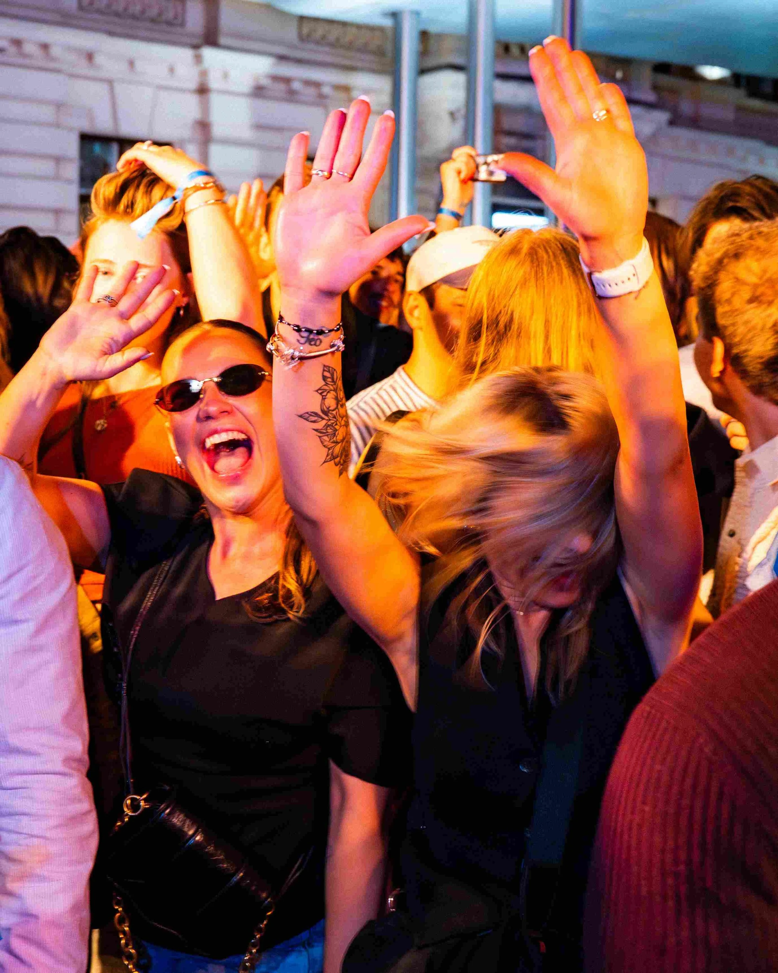 Crowd of young people at a concert or event, with two women raising their hands in the foreground, one with sunglasses and tattoos, smiling and enjoying the moment, in a lively, indoor setting.