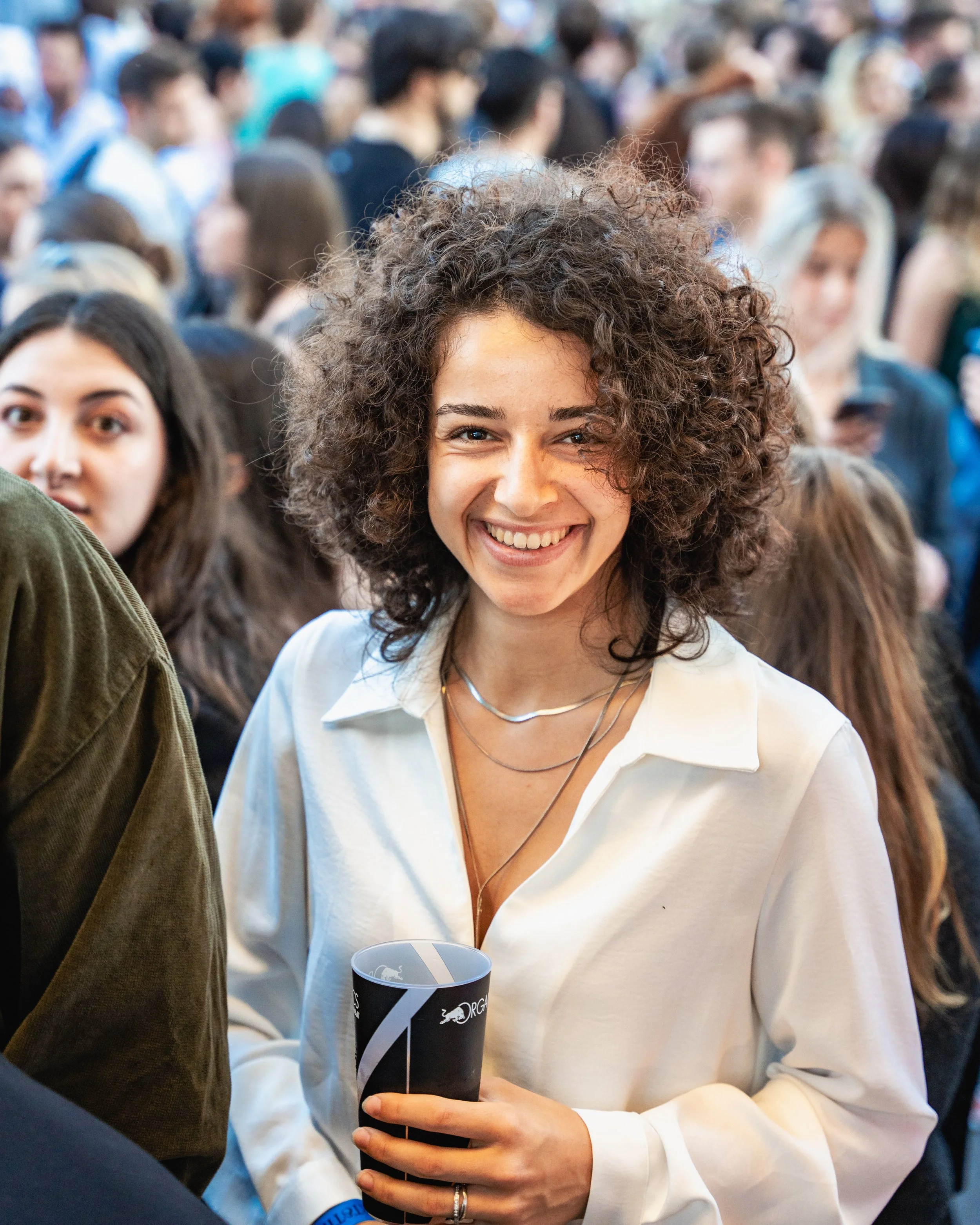 A smiling woman with curly brown hair wearing a white shirt holding a black beverage cup at a crowded event.