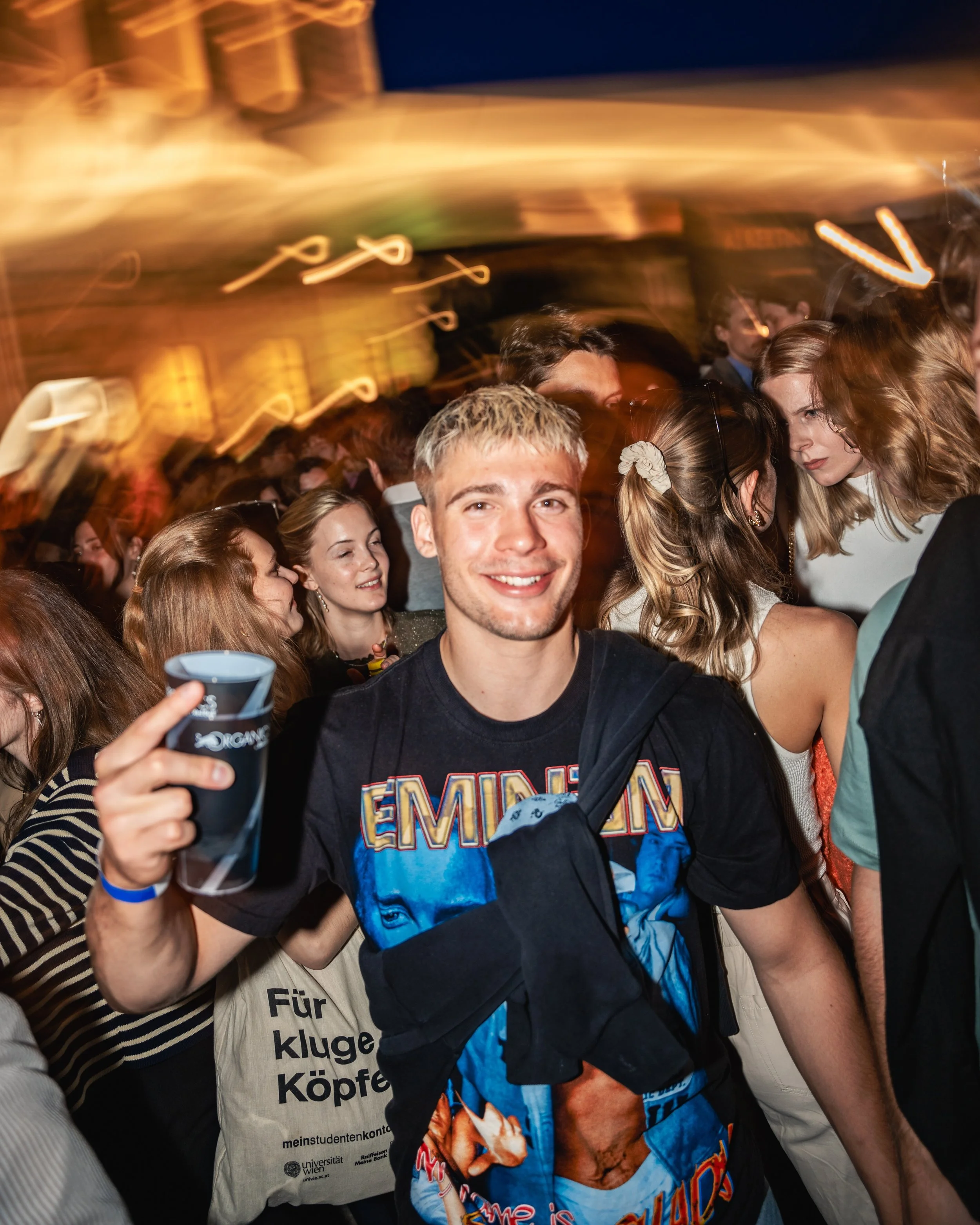 A group of young people at a party or concert, with a smiling young man in the foreground holding a cup, surrounded by others engaged in conversation, with warm and colorful lighting.