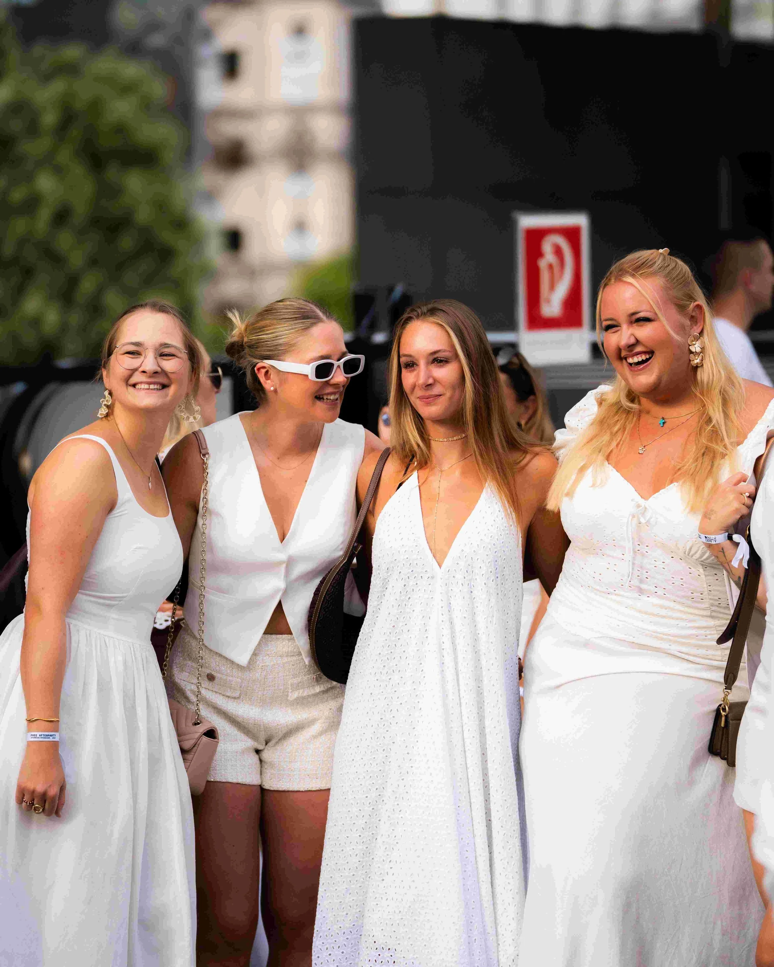 Four women standing together outdoors, smiling and chatting at a social event, with buildings in the background.