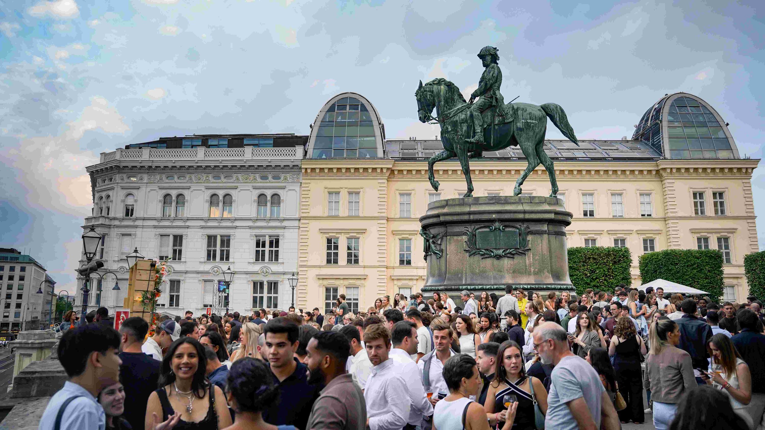 Crowd of people gathered in an urban square with historic buildings and a large equestrian statue of a man in military uniform on horseback, under a cloudy sky.