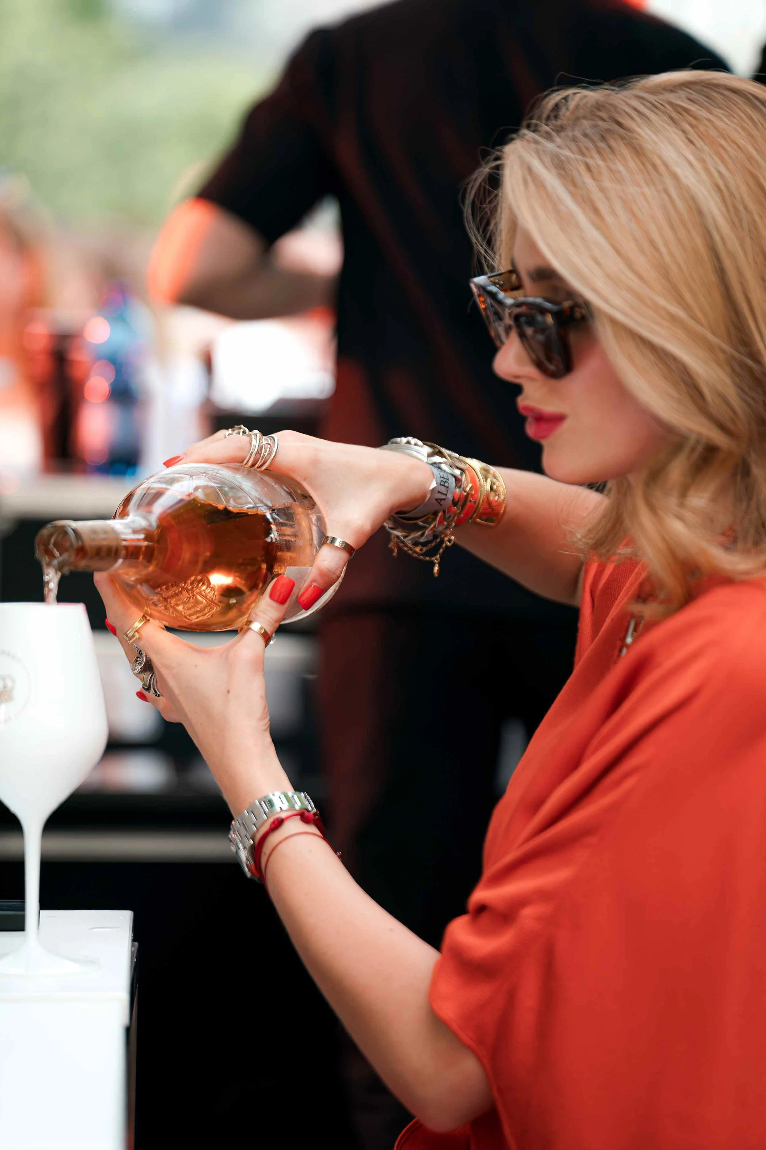 A woman with blonde hair, wearing sunglasses and bracelets, is pouring rosé wine from a bottle into a glass. She is dressed in an orange top and is outdoors, with a blurred background of people and trees.