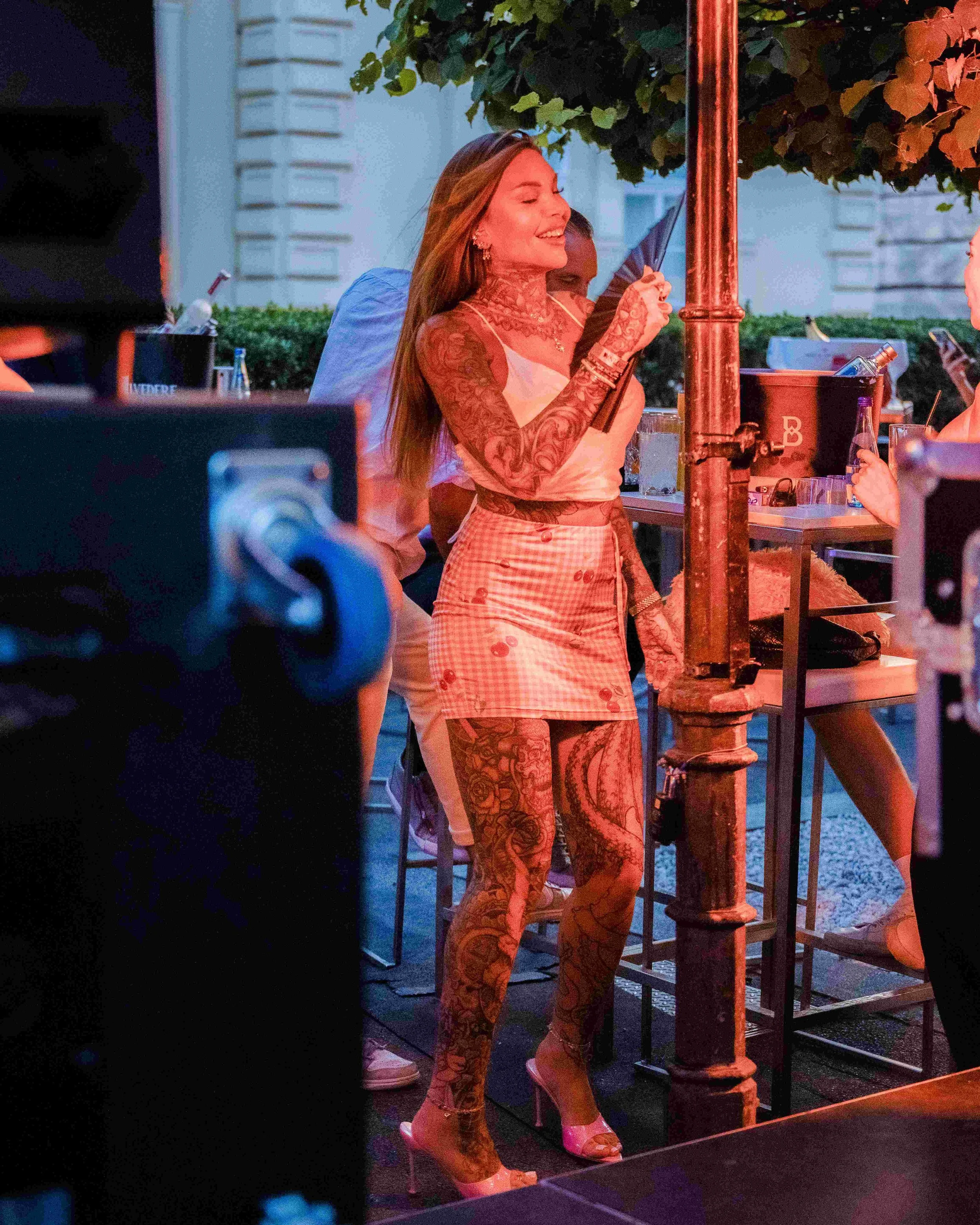 A woman in high heels and patterned clothing stands outdoors smiling, holding a black fan, with people and greenery in the background during an evening event.