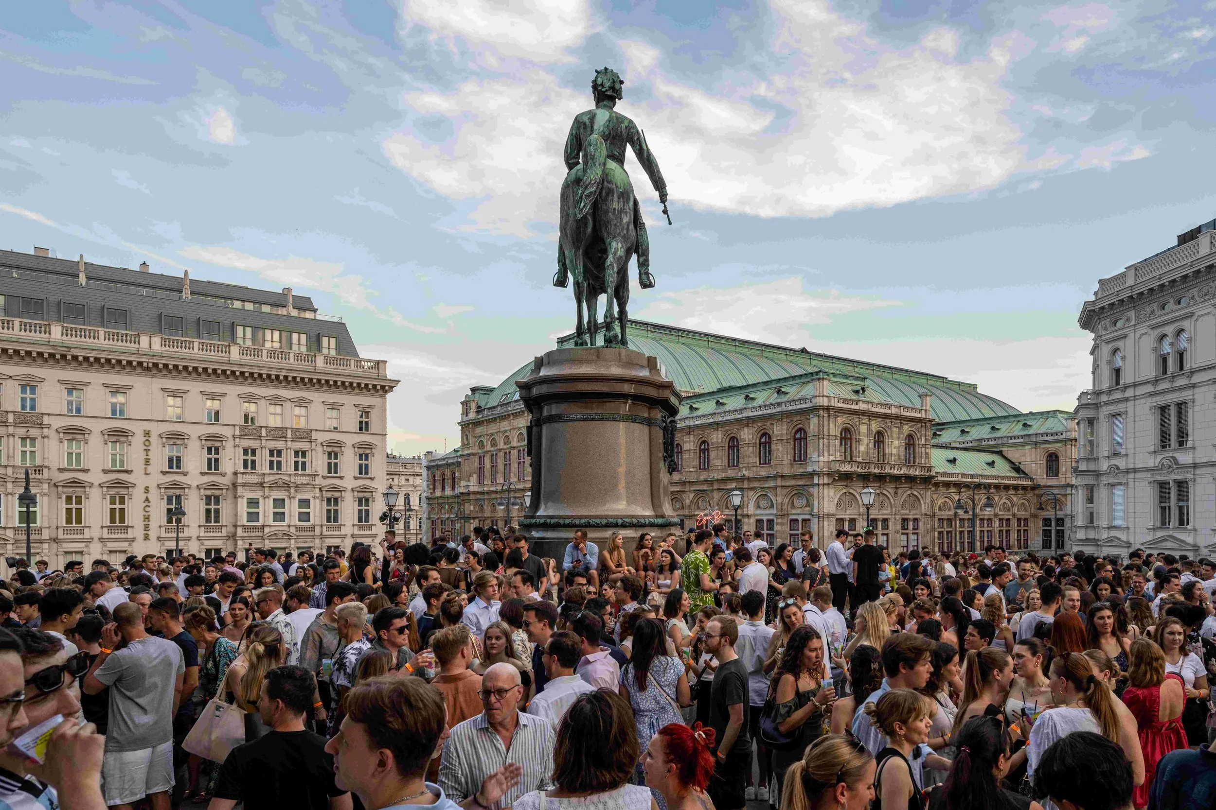 Crowd of people gathered in a city square with historic buildings, and a prominent bronze equestrian statue of a man in the background.