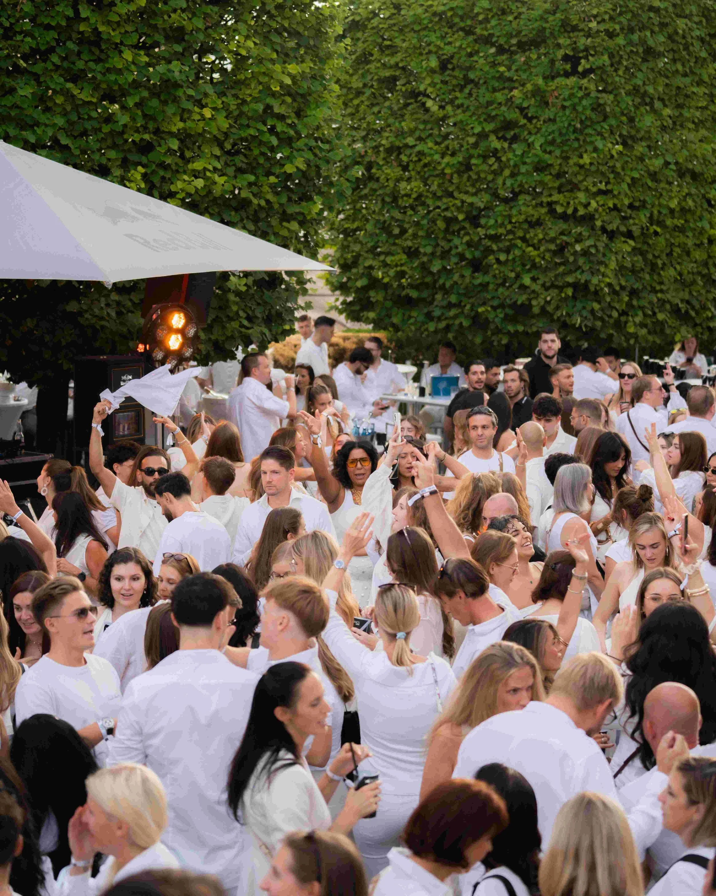 Crowd of people gathered outdoors under green trees, many wearing white shirts, some holding umbrellas, and engaging in conversation and taking photos.