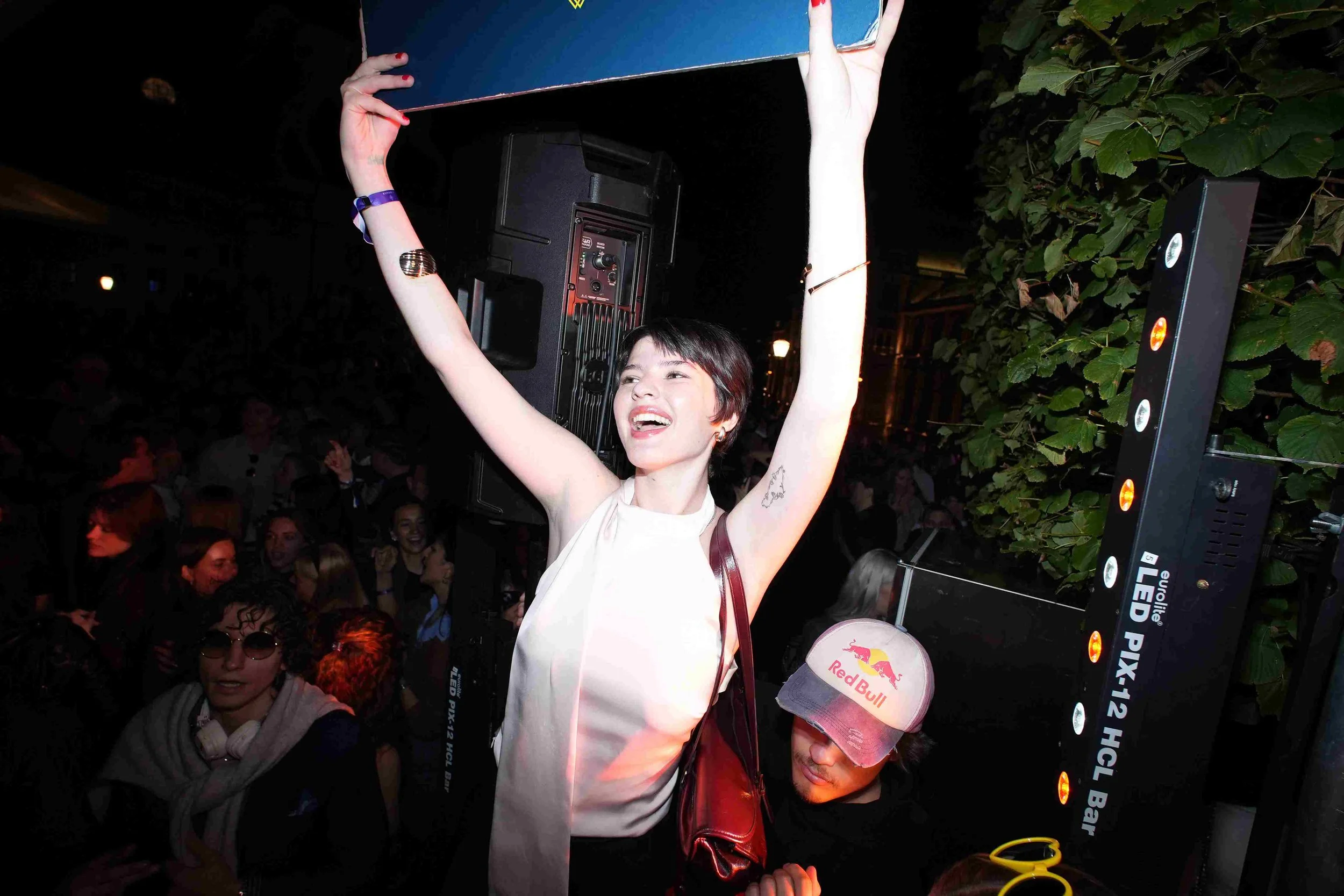 A young woman with short dark hair and tattoos on her arm is holding a large piece of equipment over her head at a crowded outdoor event at night. She is smiling and wearing a white top. In the background, there are people and some greenery, along wi
